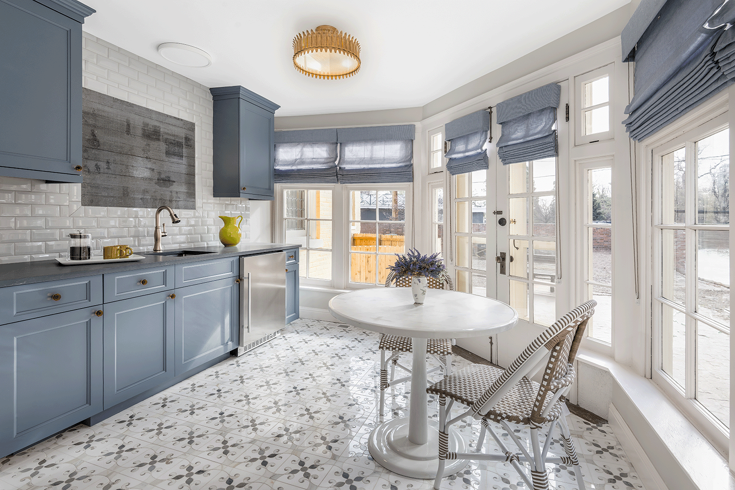 Sunroom with blue cabinets, marble mosaic tile floor, and a small round white table with two woven chairs near large windows with matching Roman shades.