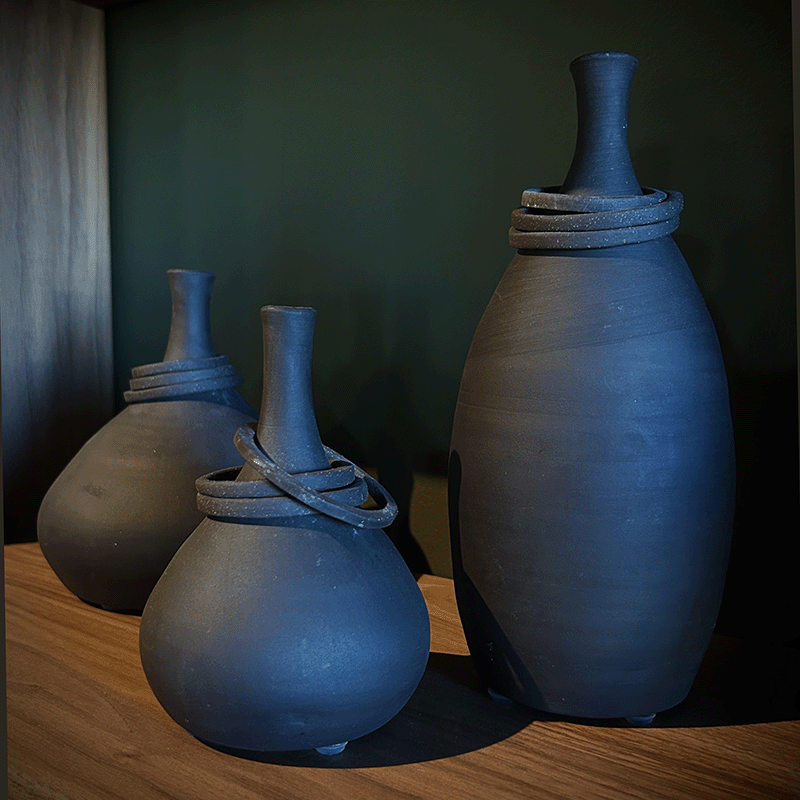Three black ceramic vases with narrow necks and coiled decoration near the top, arranged on a wooden surface against a dark green background.