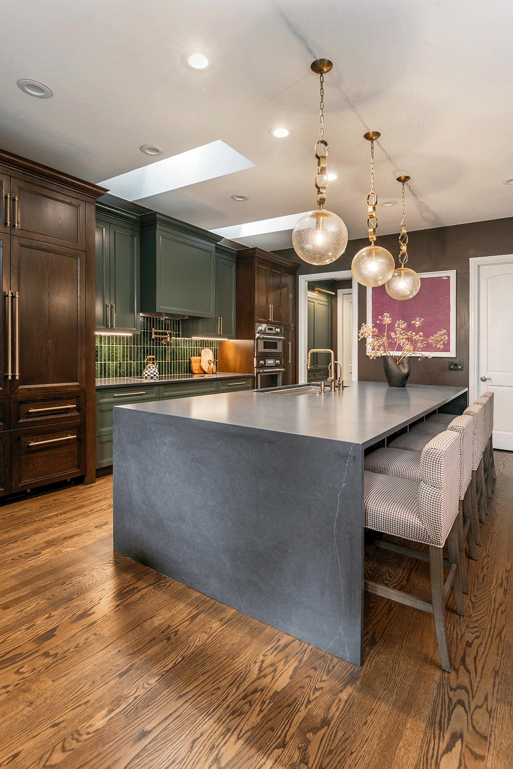 Modern kitchen with dark wood and green cabinets, a large gray island with a built-in sink, three patterned bar chairs, pendant lights, and hardwood flooring.