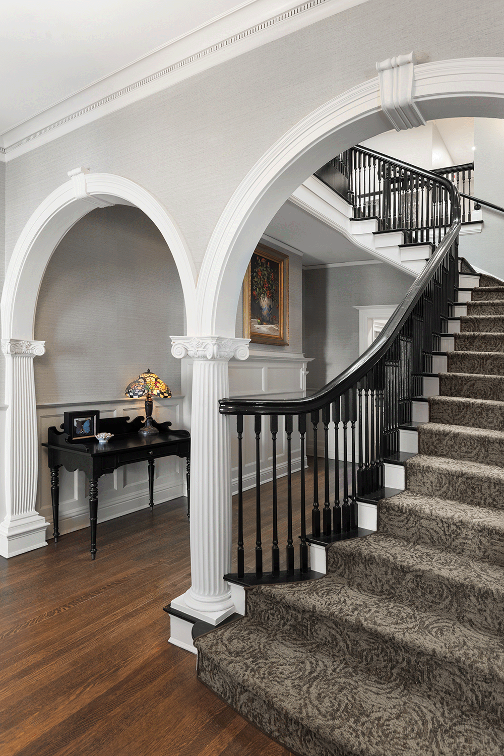 Interior of a house featuring a curved staircase with black railings, brown patterned carpet, and a small black table with a stained glass lamp and decorative items, with white decorative archways and a framed painting on the wall.