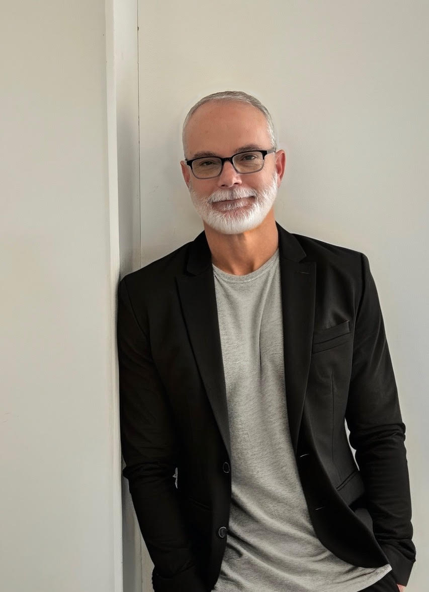 A man with gray hair, glasses, and a beard leaning against a white wall, wearing a black blazer over a gray t-shirt.