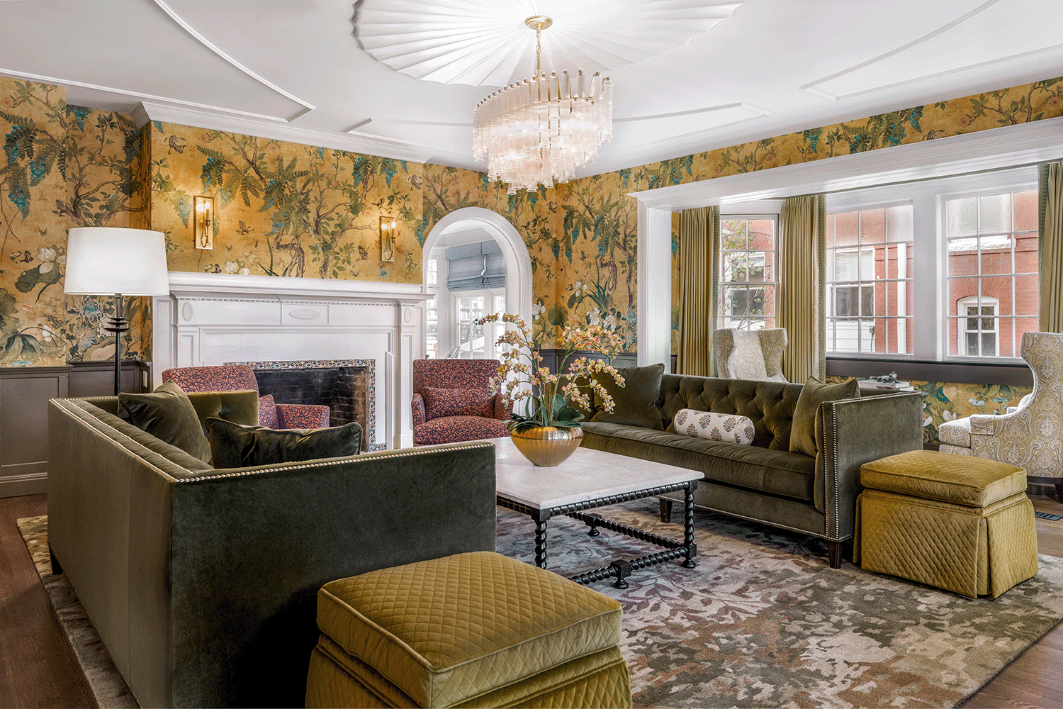 A cozy living room with floral wallpaper, large windows with yellow curtains, a white fireplace, and a variety of upholstered chairs and sofas around a central coffee table with a decorative plant. There is a chandelier hanging from the ceiling.