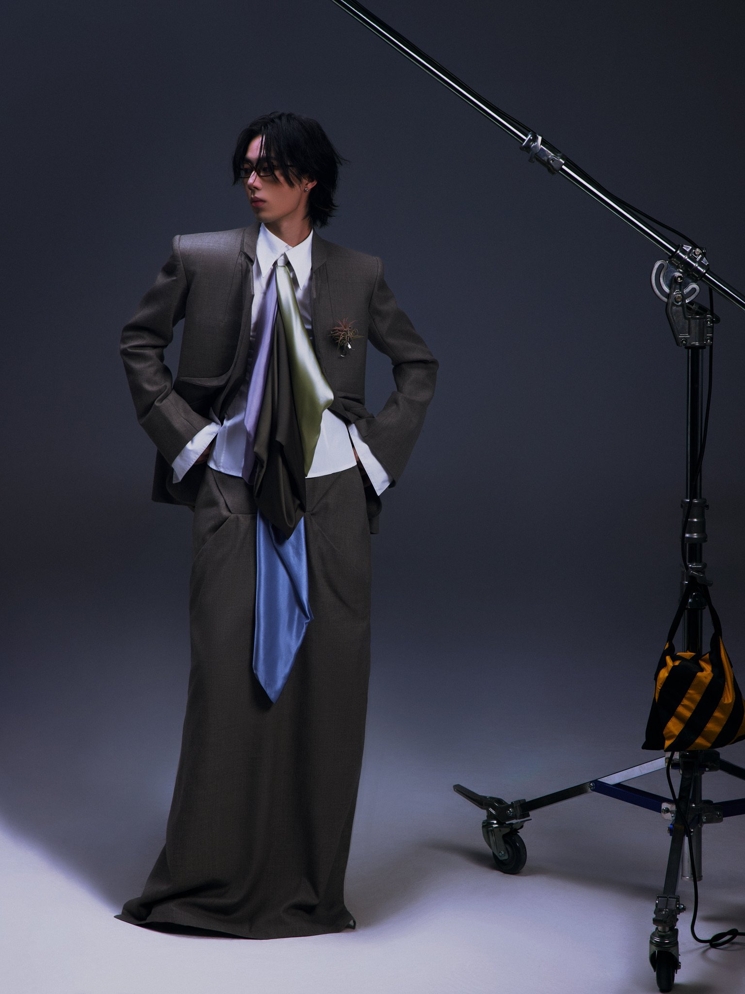 Person with black hair and glasses wearing a gray suit and tie, standing in a studio with a dark background, next to a large photography light stand.
