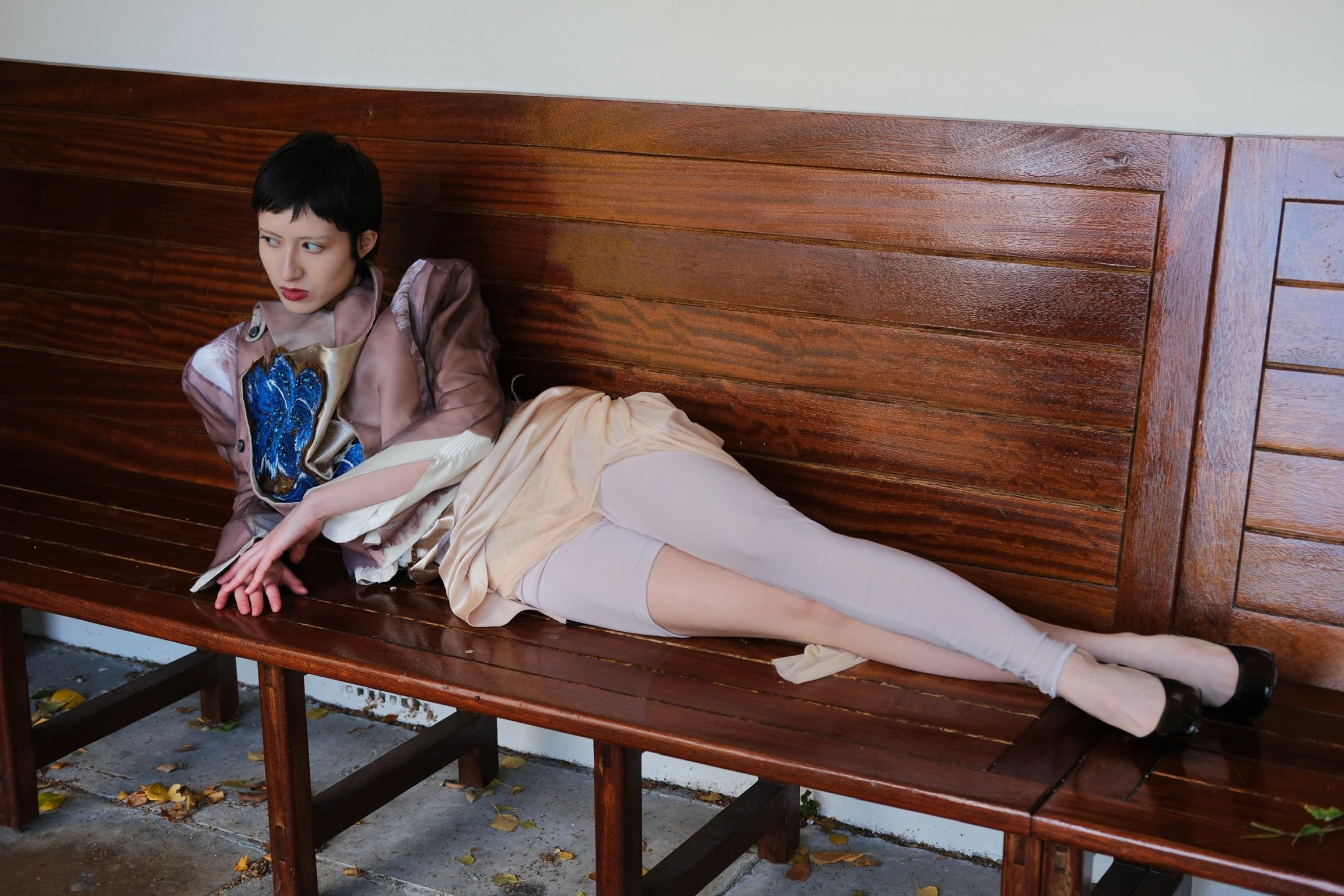 A person with short black hair and red lipstick lying on a wooden bench outdoors, wearing a patterned satin jacket, a beige skirt, white tights, and black shoes, with a neutral expression and looking to the side.