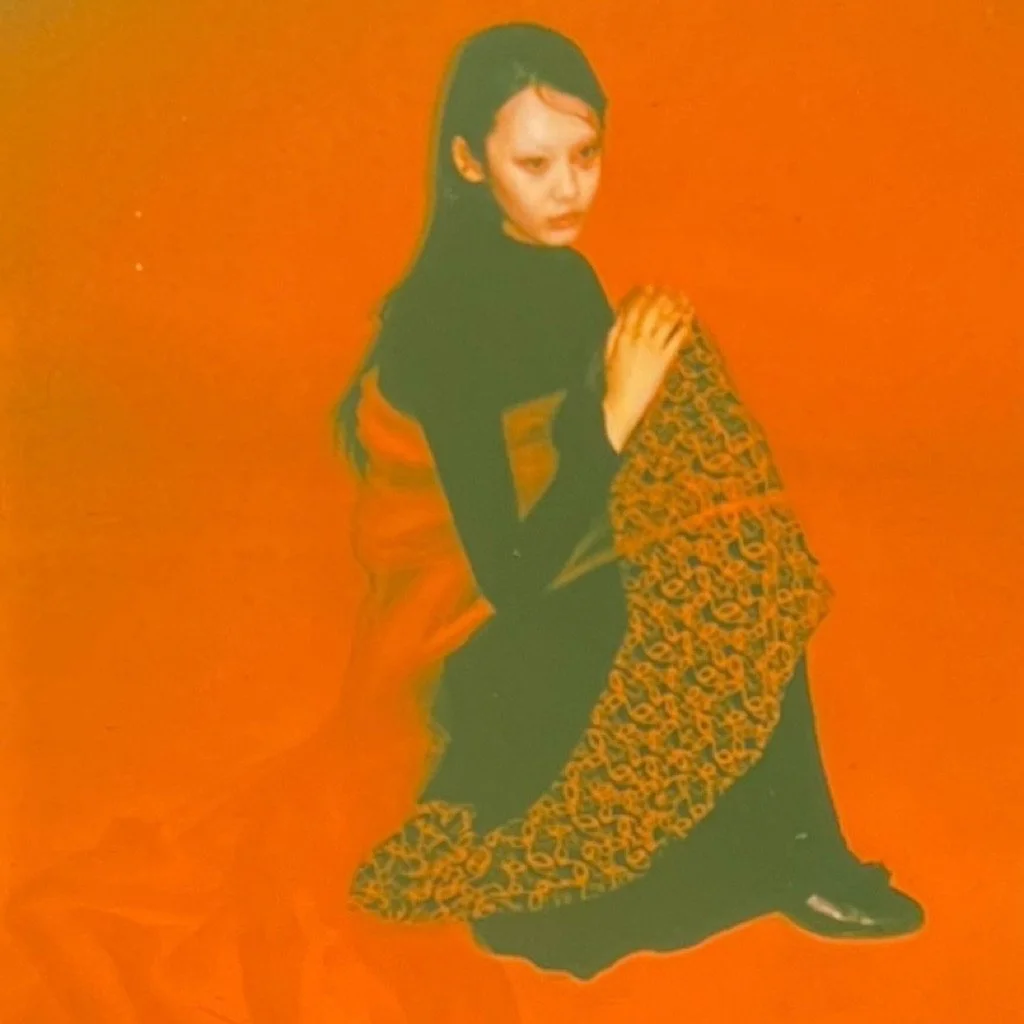 A young woman with black hair and light makeup sits against a bright orange background, wearing a black top and a colorful patterned skirt, looking slightly to the side.