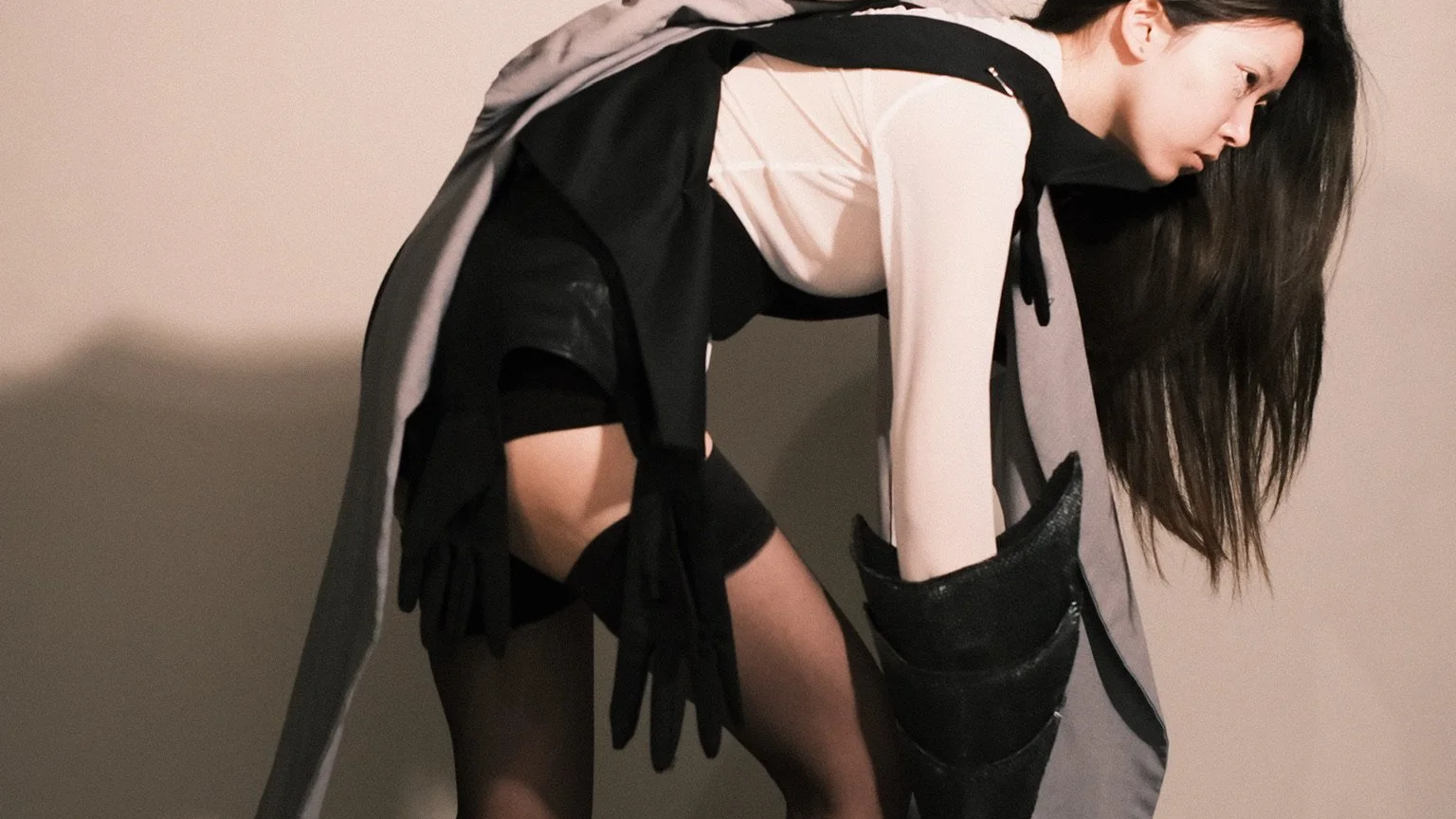 A woman in black gloves, thigh-high stockings, and a black dress is bent over, leaning forward with long dark hair covering part of her face, against a plain wall.