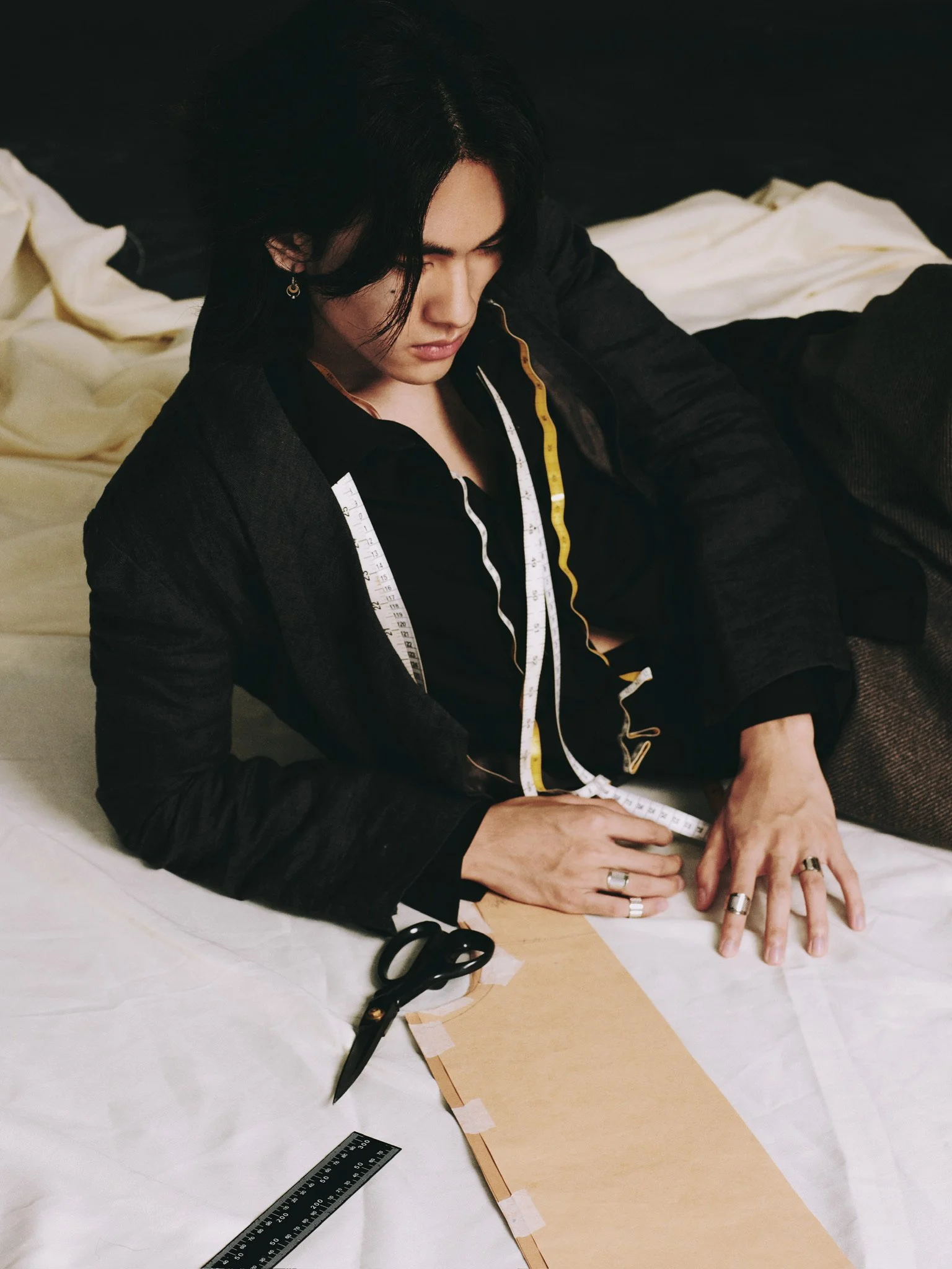 A person with black hair measuring fabric with a tape measure, sitting at a table with scissors and fabric pieces.