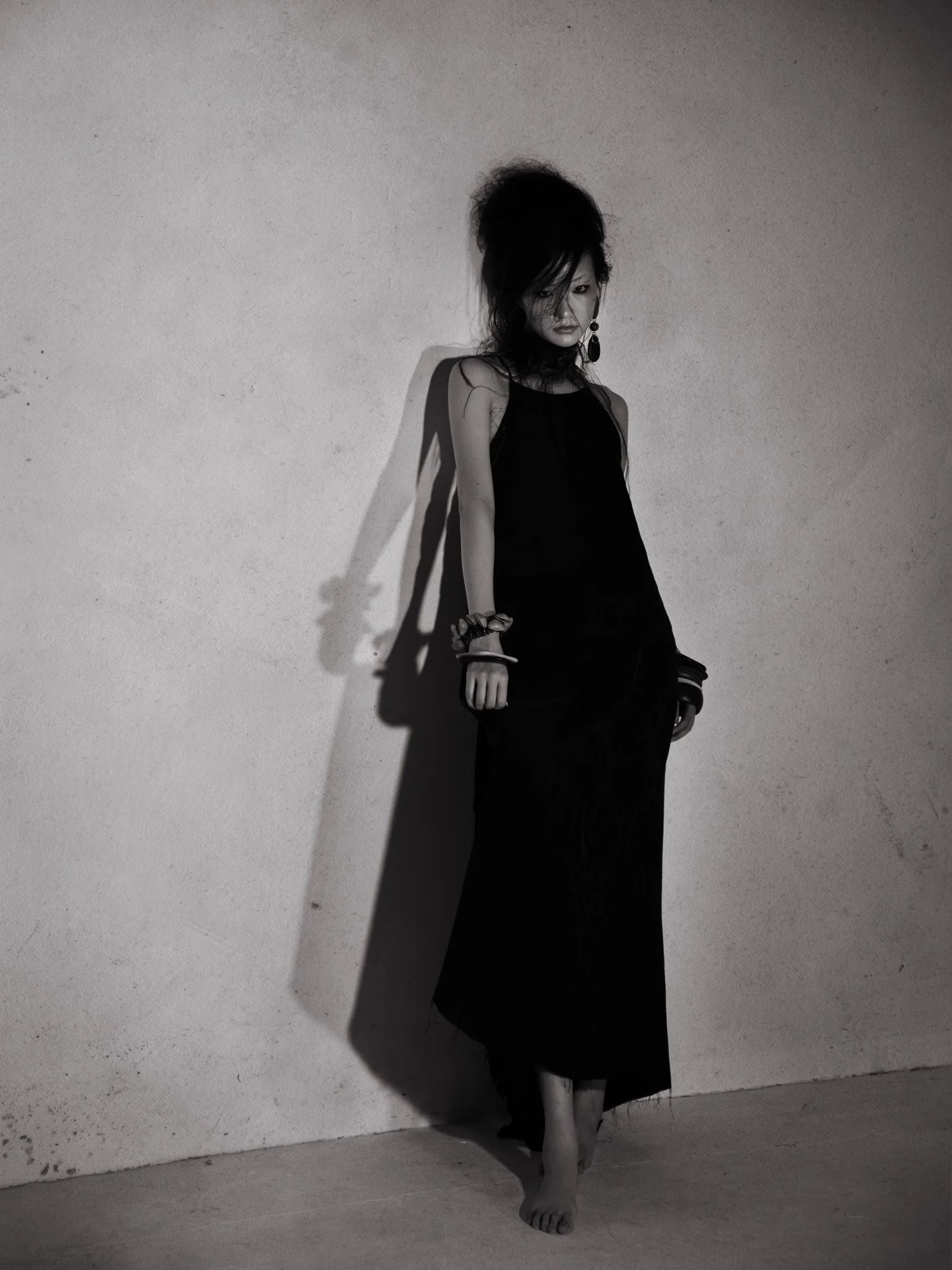 A black and white photo of a woman in a long, dark dress, standing against a plain wall, with her hair styled in a voluminous updo, wearing large earrings, and striking a contemplative pose.