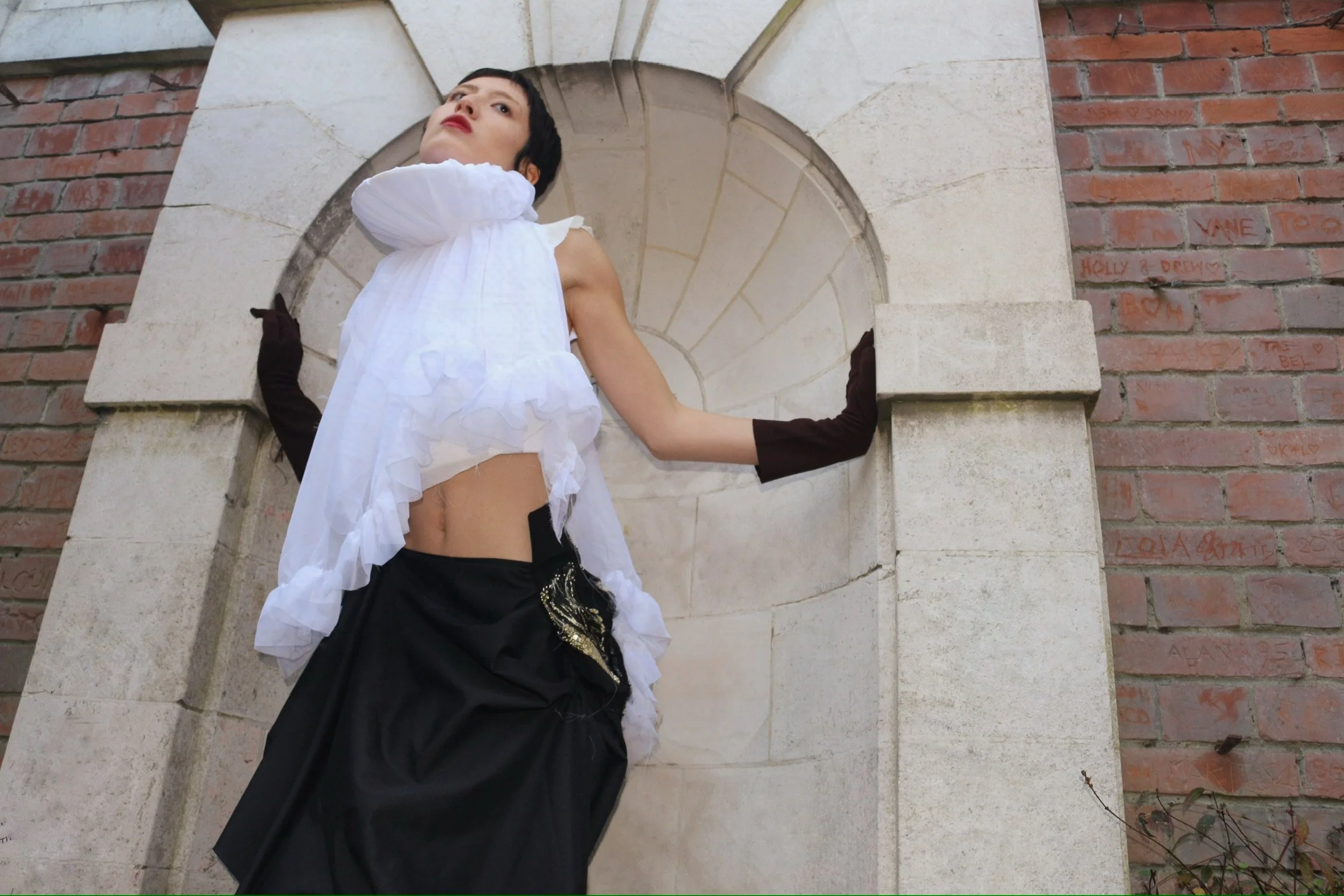 A woman with short black hair and red lipstick standing in front of a stone archway with brick walls on either side, wearing a white sleeveless top, black pants, and long black gloves.