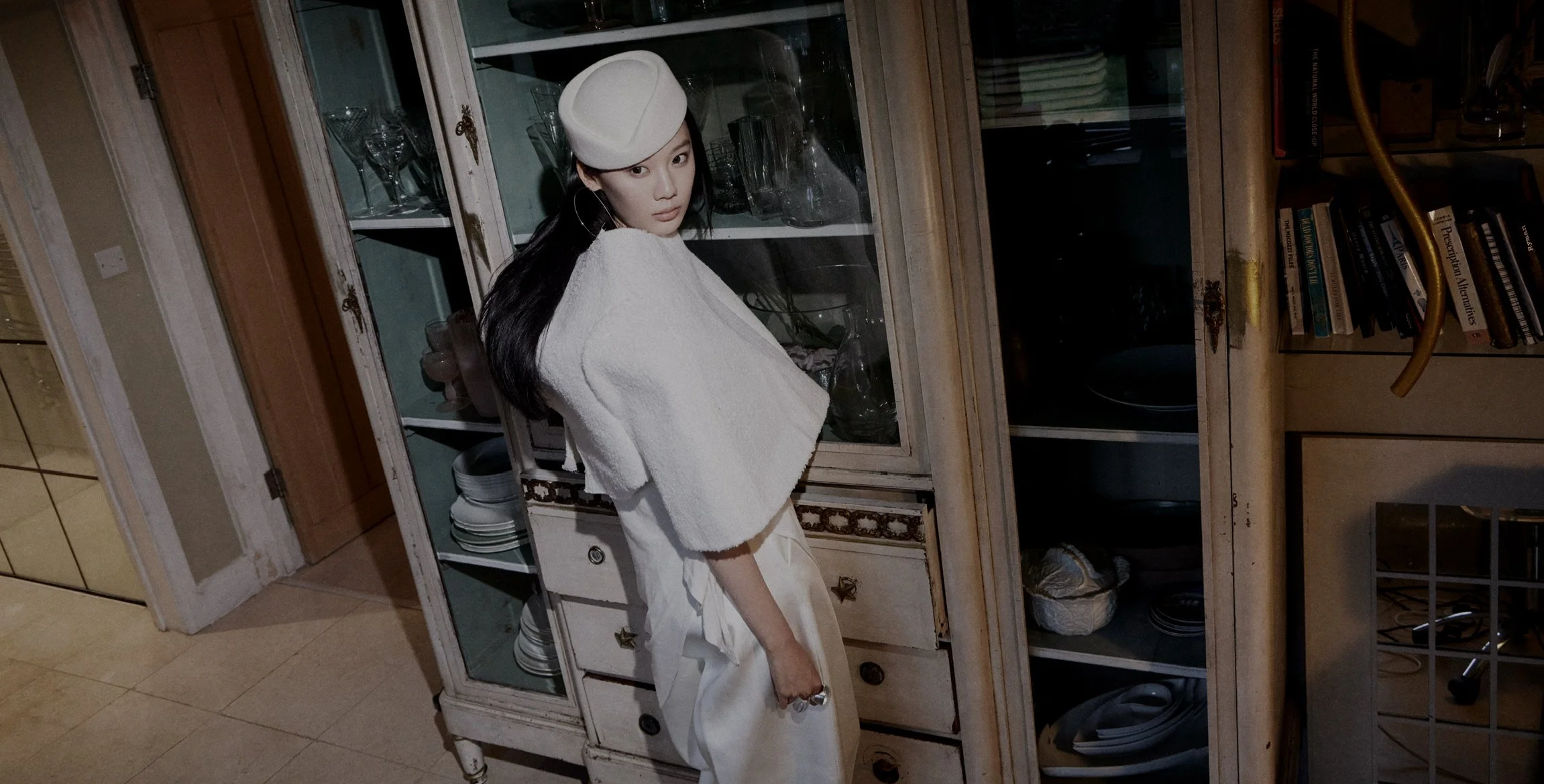A woman with long dark hair wearing a white hat, white bathrobe, and large hoop earrings standing in a room with a vintage glass-fronted cabinet filled with glassware and dishes, and a bookshelf with books visible in the background.