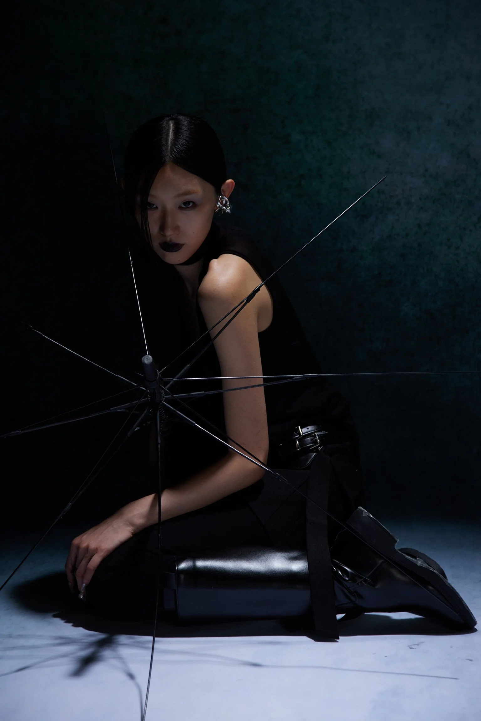 A woman with dark hair and dark lipstick posing against a dark background, sitting on the floor with her arm resting on her knee, surrounded by black umbrellas.