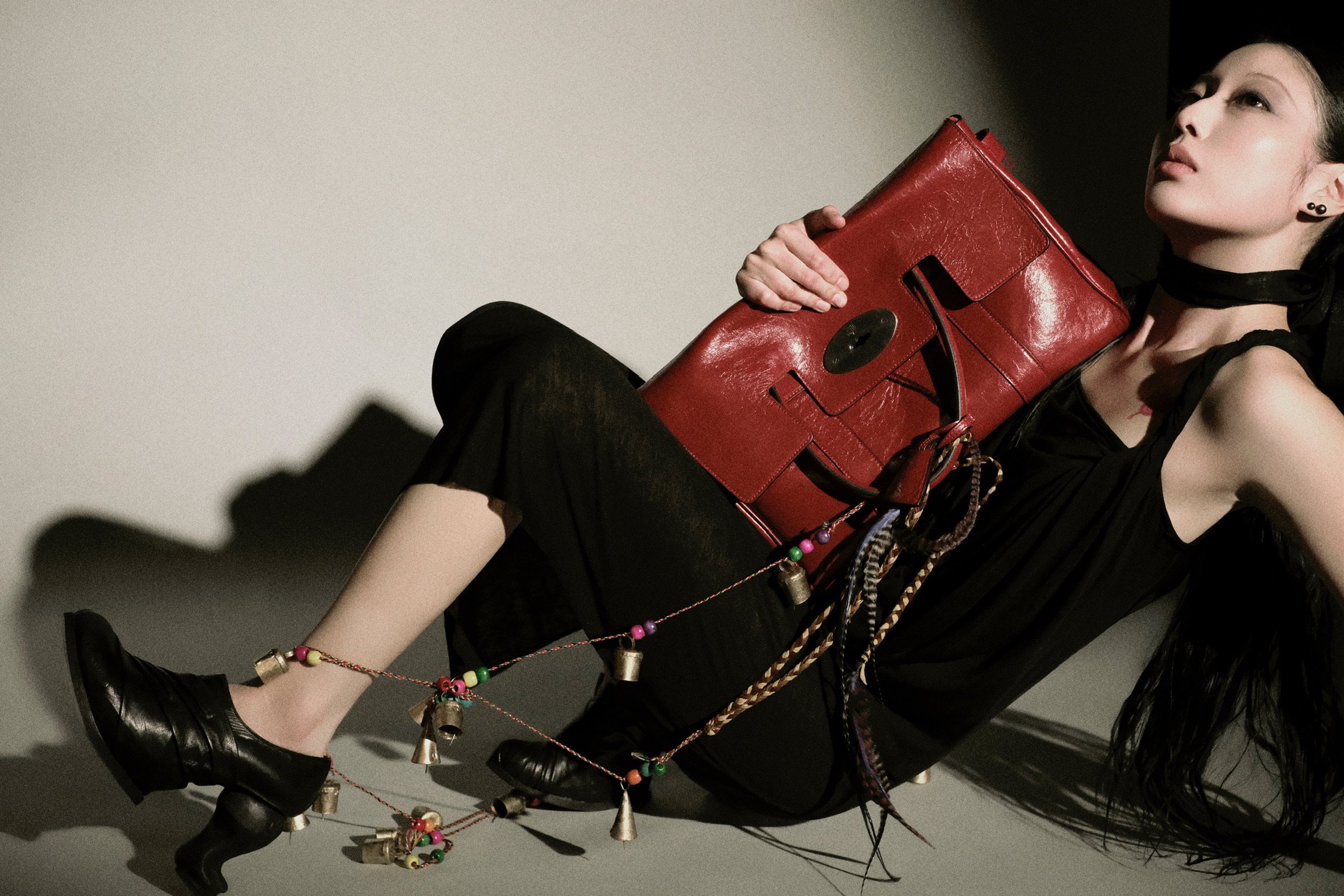 A young woman with dark hair, styled in waves, wearing a black sleeveless top, black shorts, black boots, and black earrings, holding a large red handbag. She is reclining against a light-colored wall with shadows cast nearby.
