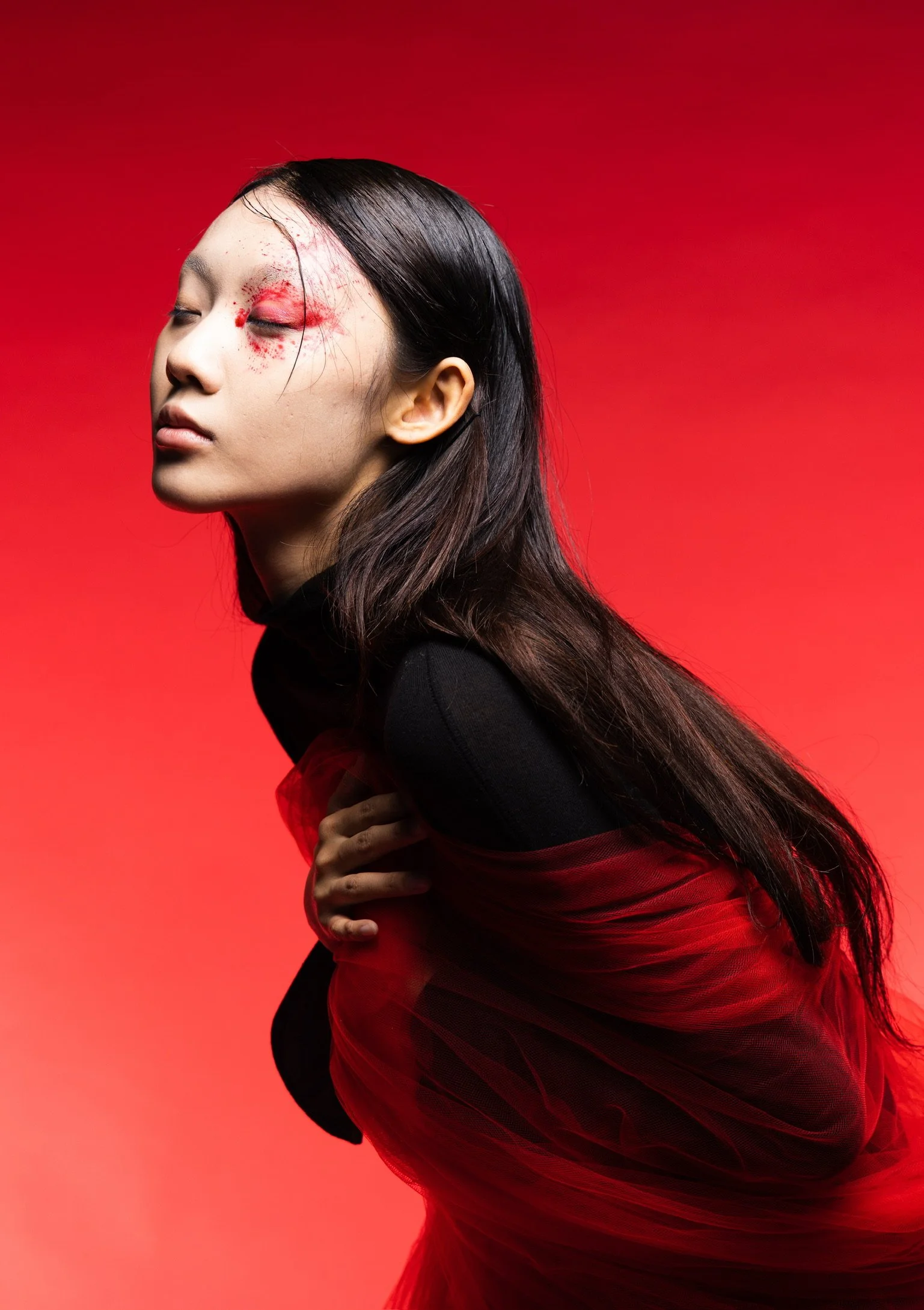 A woman with long dark hair and closed eyes, wearing a black and red outfit, poses against a red background with artistic makeup around her eye, creating a bold and creative visual effect.