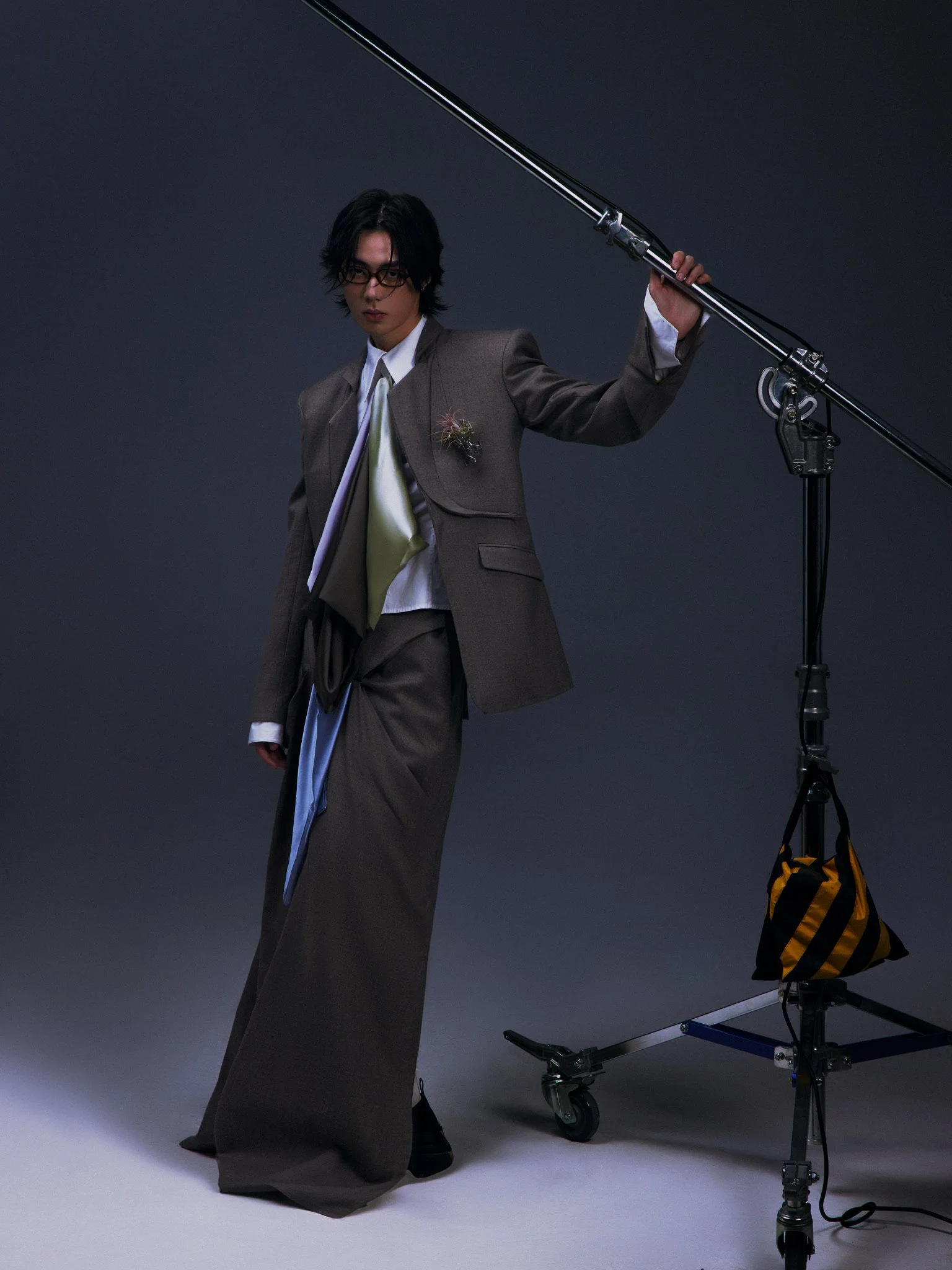 A young man with black hair and glasses dressed in a gray suit and loose tie, standing in a photo studio, holding a large camera boom arm with a light stand and equipment nearby.