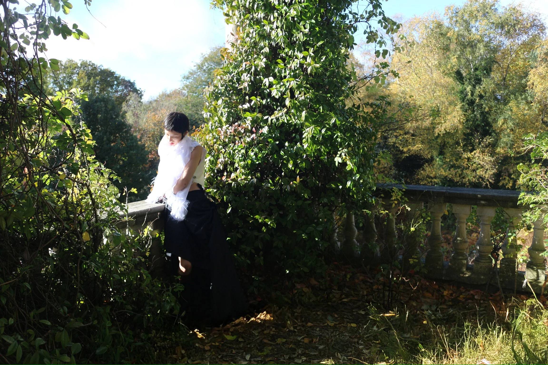 A person with short dark hair wearing a white top and a black skirt, standing outdoors in front of a green bush and trees, with sunlight filtering through the foliage.