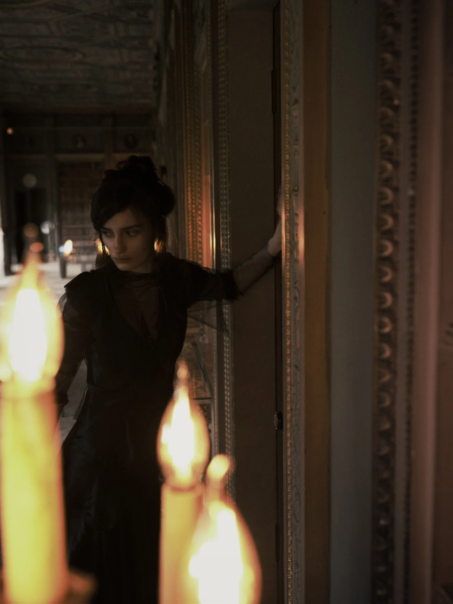 A woman with dark hair styled in a messy bun, wearing dark clothing, stands near a mirrored wall in a dimly lit room with ornate gold and patterned moldings. In the foreground, there are blurred candle flames, and the woman appears to be gazing downward.