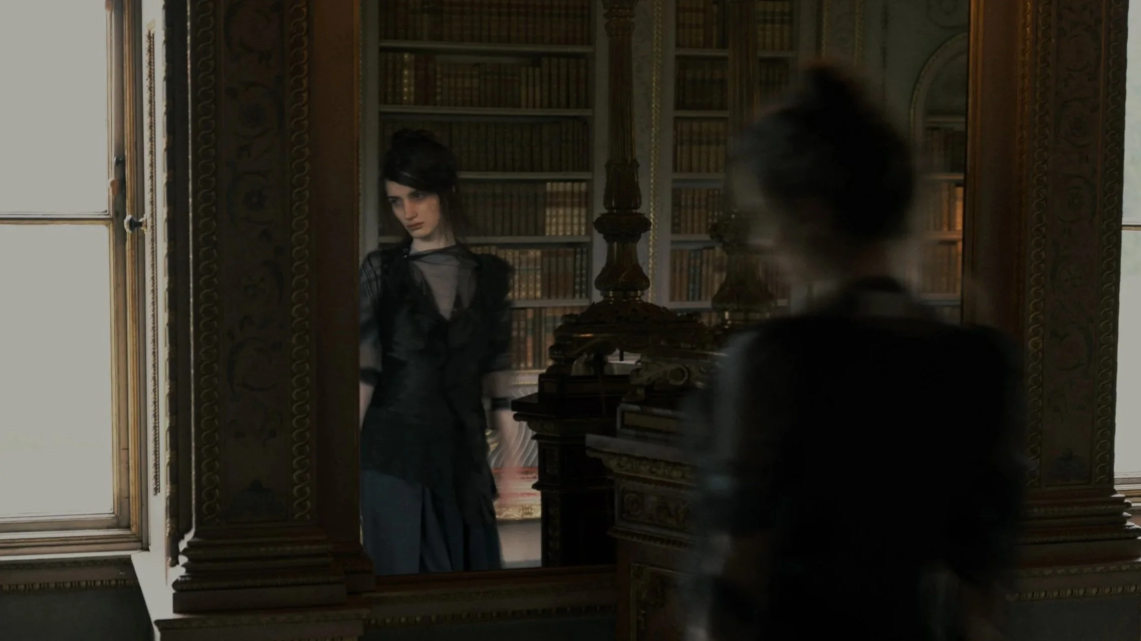 A woman with dark hair wearing a sheer black top and black skirt looking at her reflection in a mirror in a room with ornate woodwork and a bookshelf filled with books.