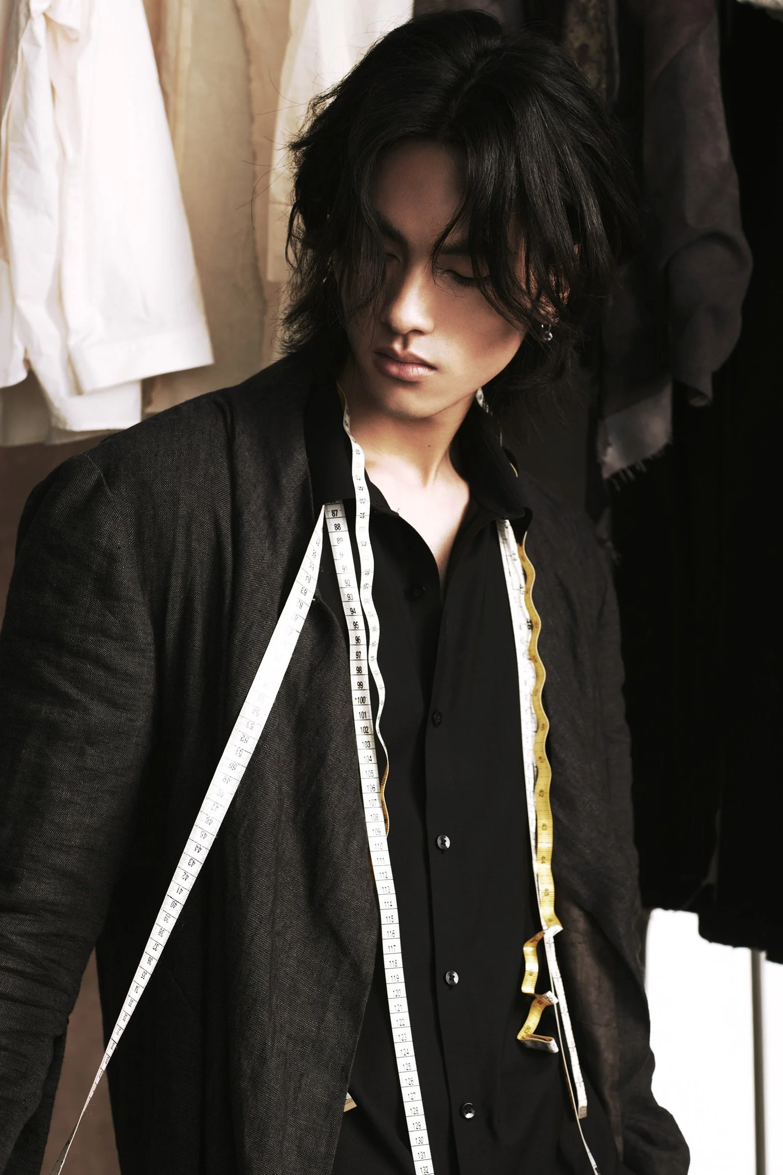 A woman with shoulder-length dark hair in a black blazer, with a measuring tape draped around her neck, in a room with hanging clothes in the background.