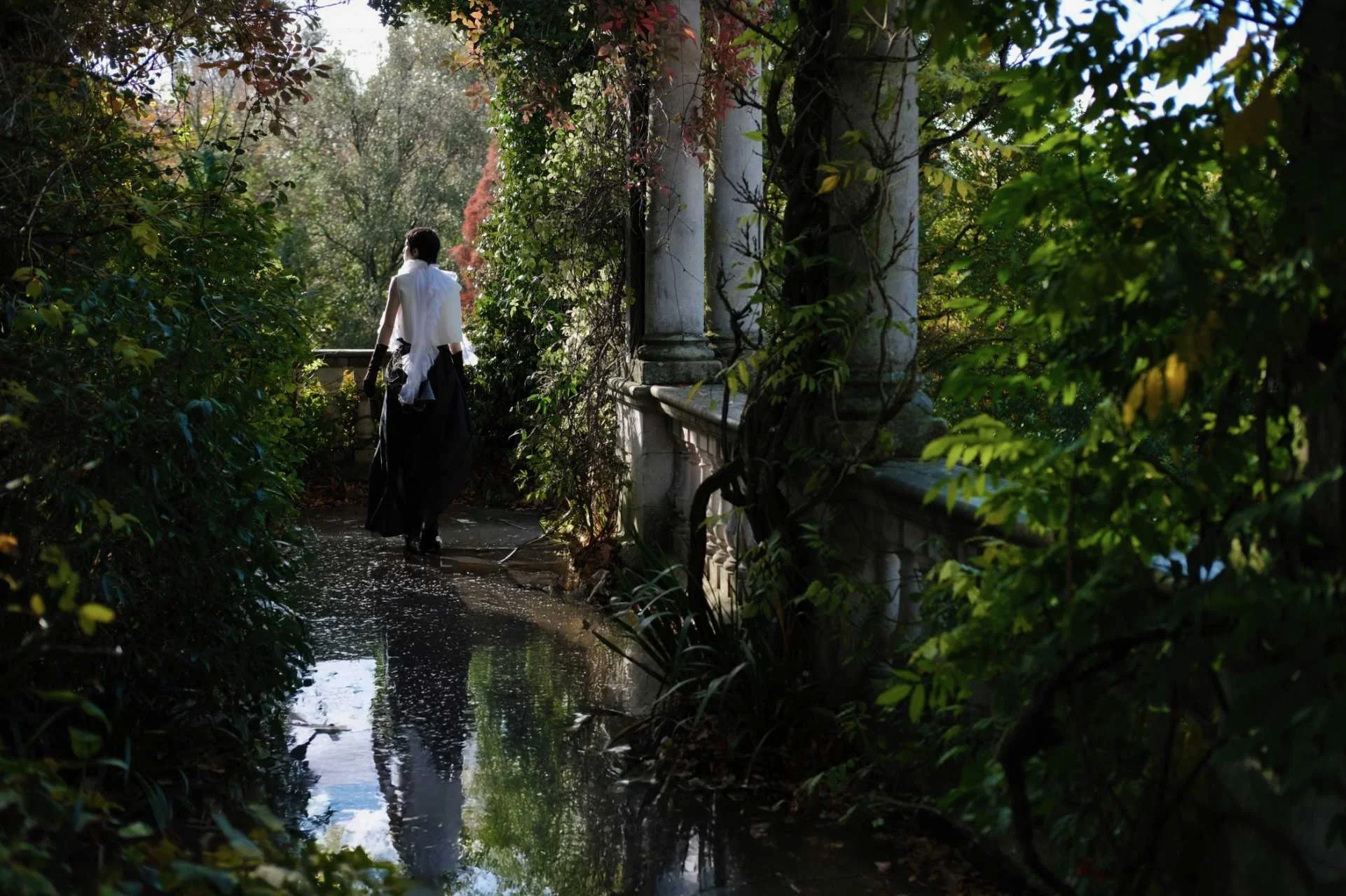 A person dressed in black and white walking through a lush, green garden with a small waterway reflecting trees and the sky.