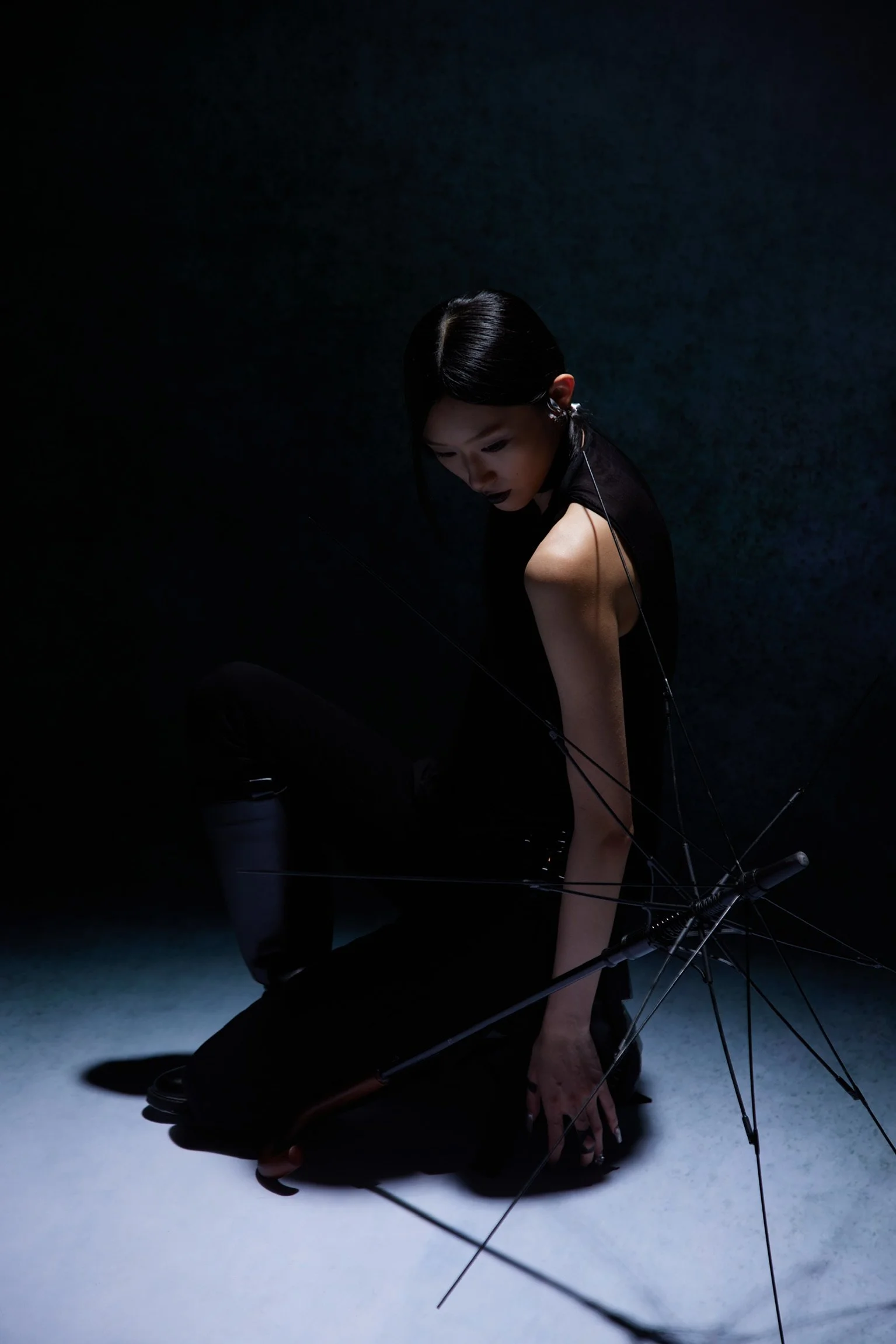 A woman with black hair and dark makeup, dressed in black, leaning forward on the ground with her hand on a white surface. An open umbrella lies on the ground beside her, surrounded by thin, black rods. The lighting creates a dramatic effect, illuminating her face and arm against a dark background.