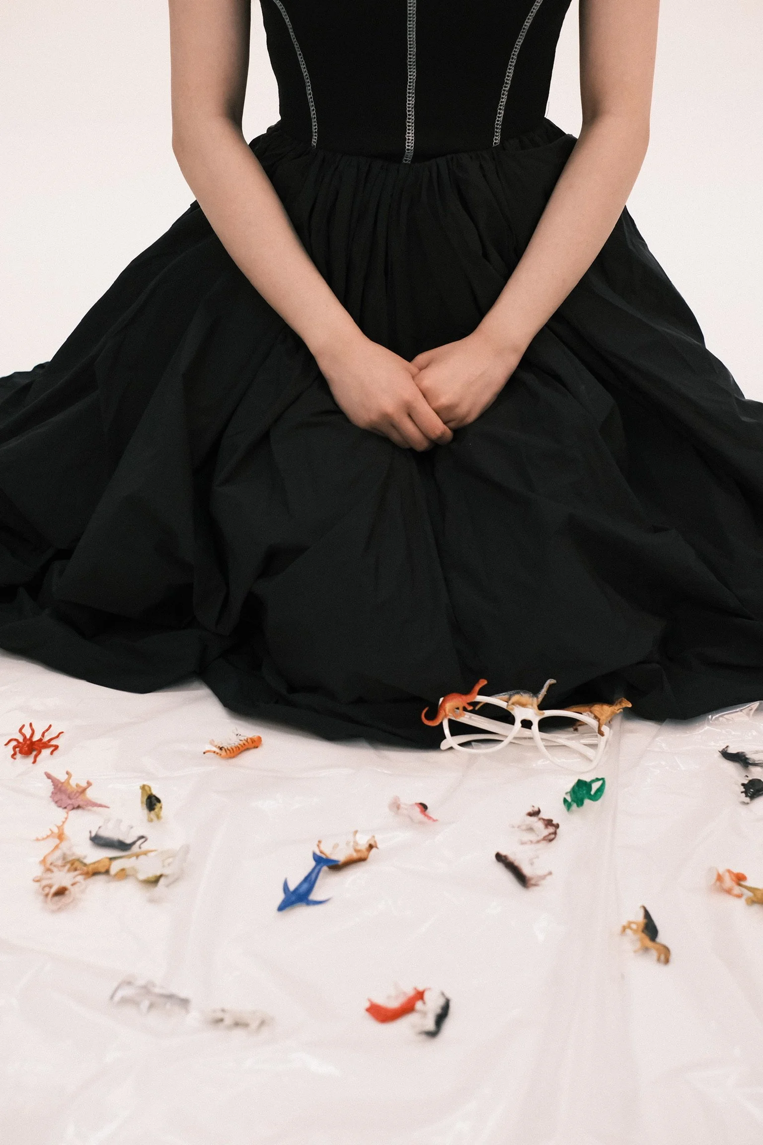 Person wearing a black dress sitting with hands clasped, surrounded by scattered small plastic toy animals and sunglasses on a white surface.