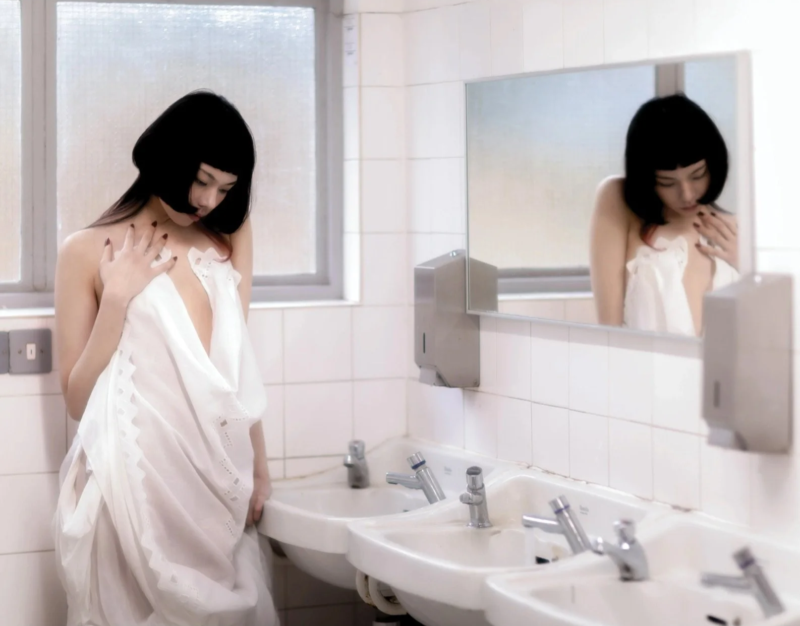 A woman with black hair and wearing a white dress standing in front of a bathroom sink, looking down, with a mirror reflecting her image. The bathroom has white tiled walls and three sinks with silver faucets.