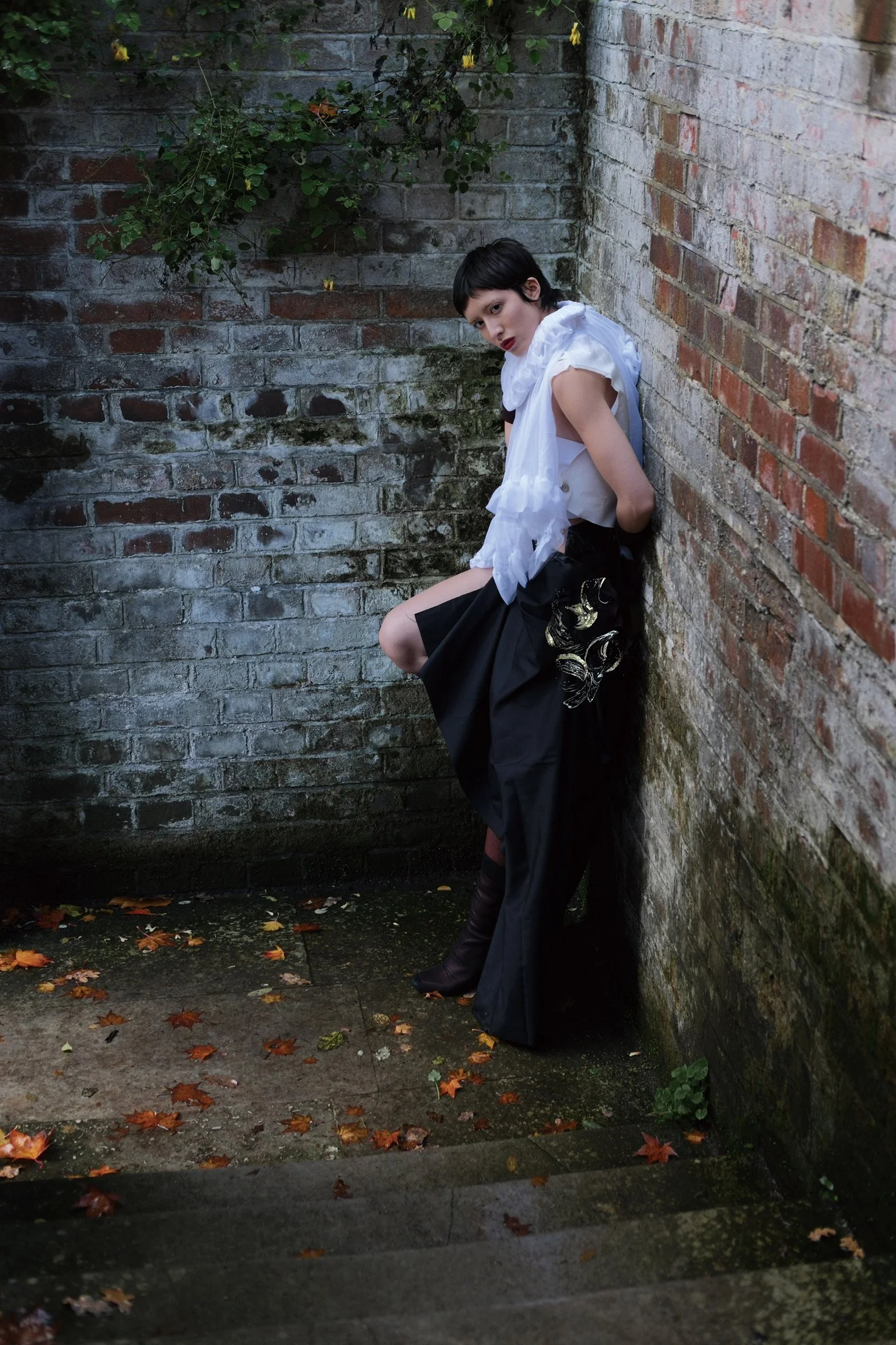 A person leaning against a brick wall in an outdoor setting with fallen autumn leaves on the ground.