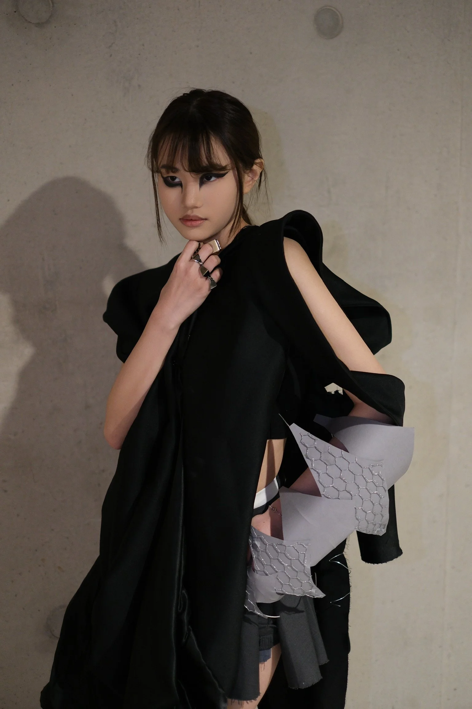 Fashion-forward woman with bold eye makeup and layered black clothing, standing against a plain wall with shadows.