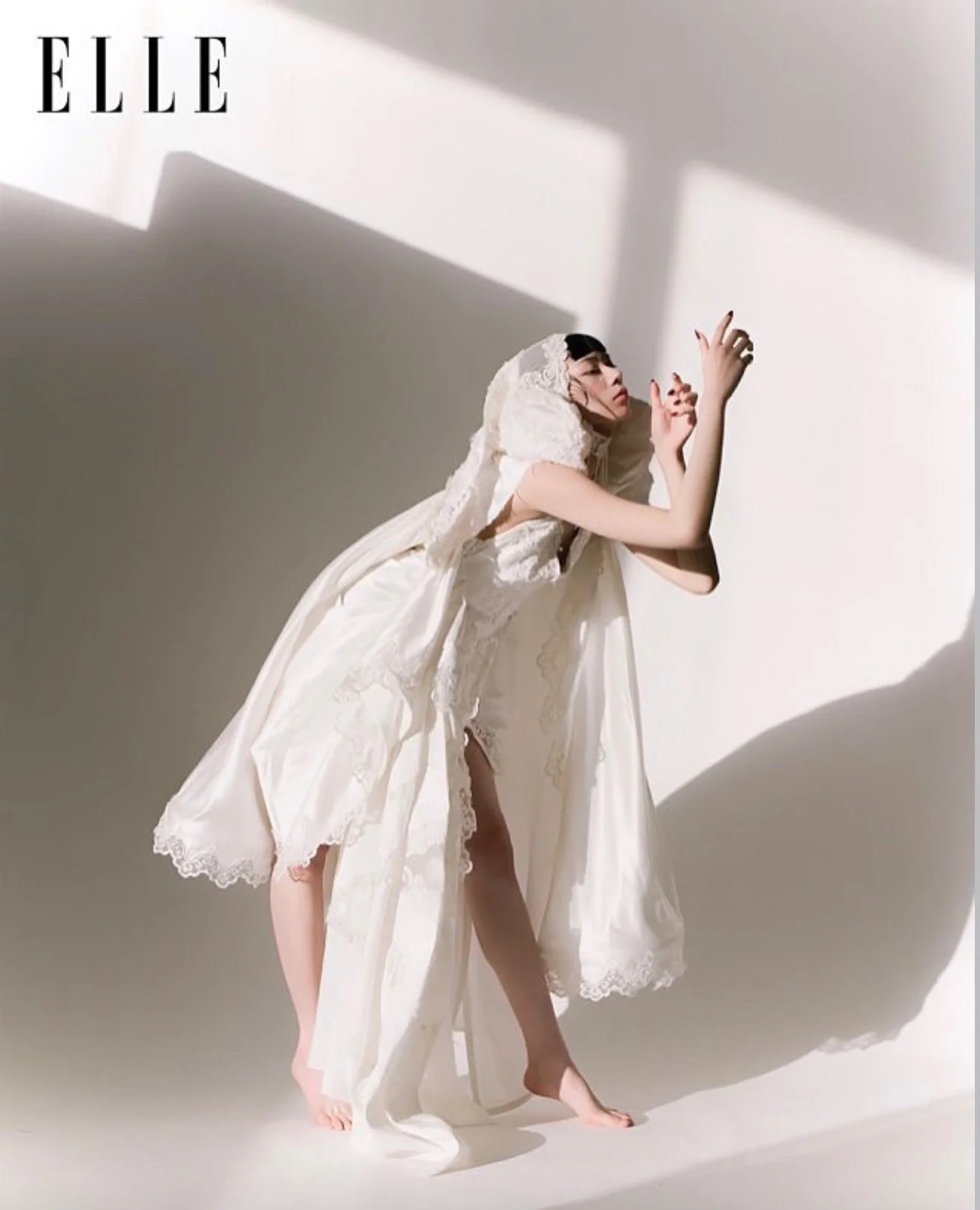 A model wearing a vintage white lace dress with a hood, posing against a plain backdrop with shadows cast on the wall.