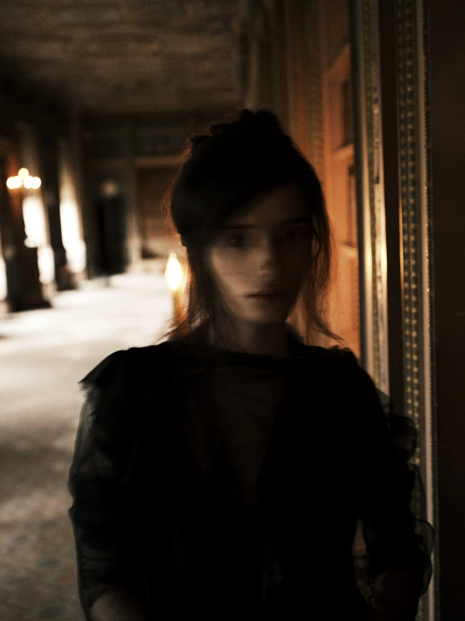 A woman with dark hair and a bun hairstyles, wearing a dark jacket and standing in a dimly lit corridor with ornate wallpaper and lamps in the background.