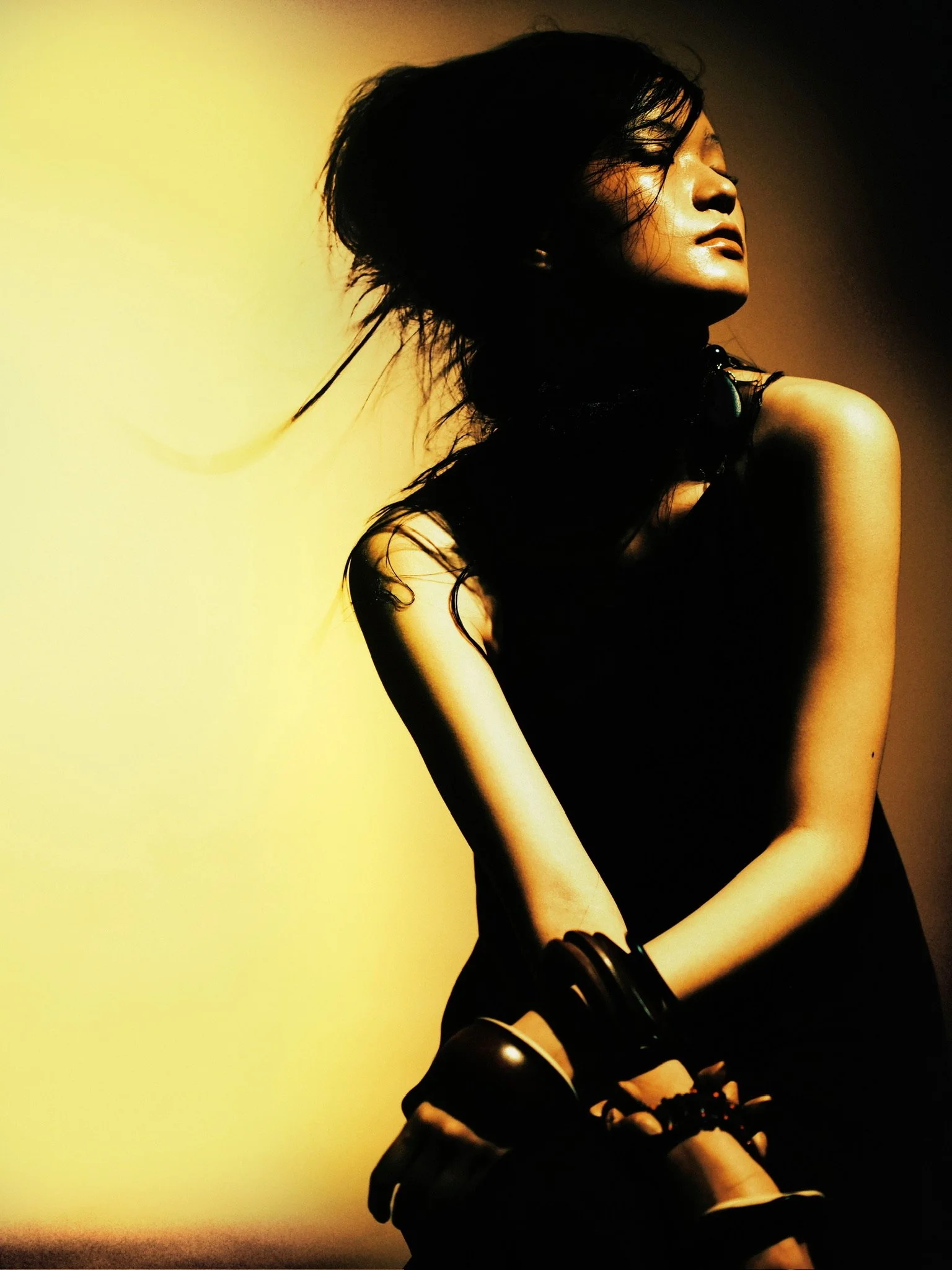 A woman with long dark hair, wearing a black sleeveless top, sitting with her eyes closed and head tilted back, against a yellow and dark brown background, with a somber or contemplative expression.