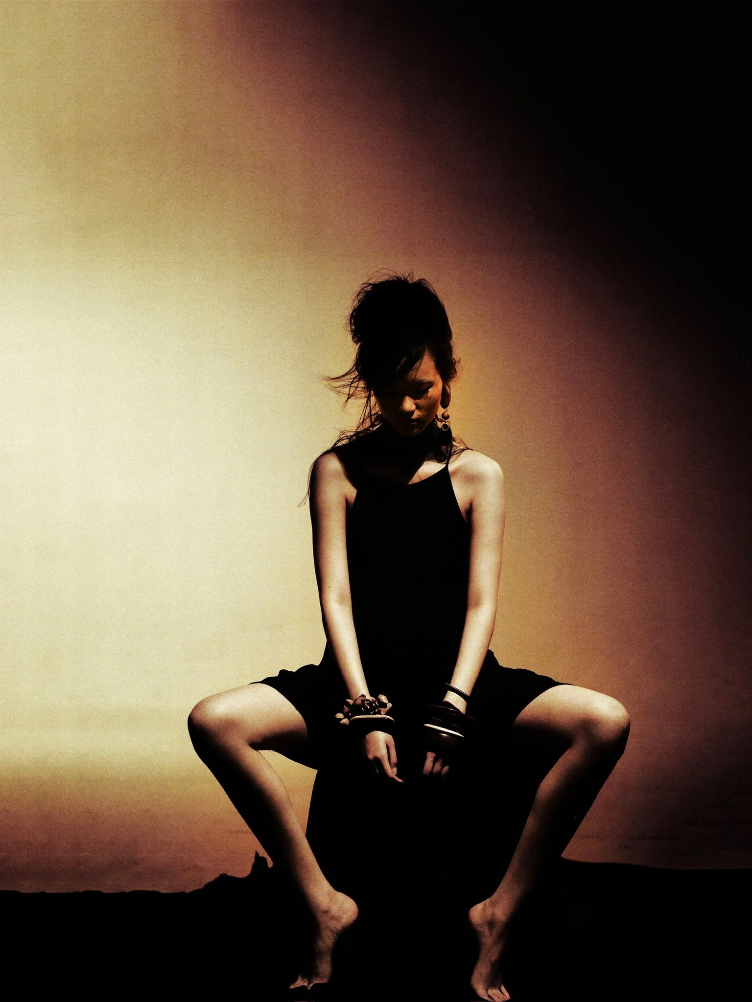 A woman sitting cross-legged on the ground with a sunset background, wearing a black dress and accessorized with earrings and bracelets.