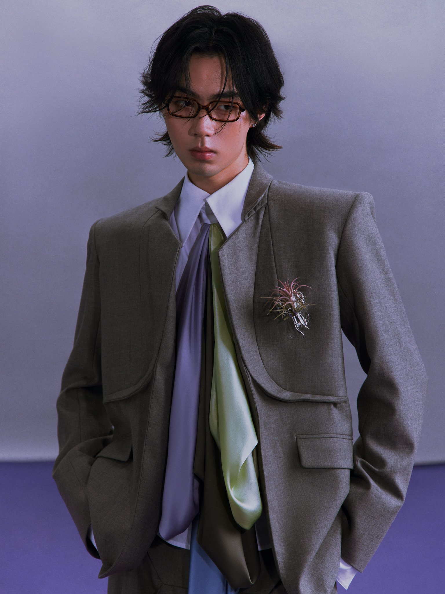A person with medium-length dark hair, wearing glasses, a gray blazer, a white shirt, and a lavender tie. They have a flower pinned to their blazer and are standing against a plain background.