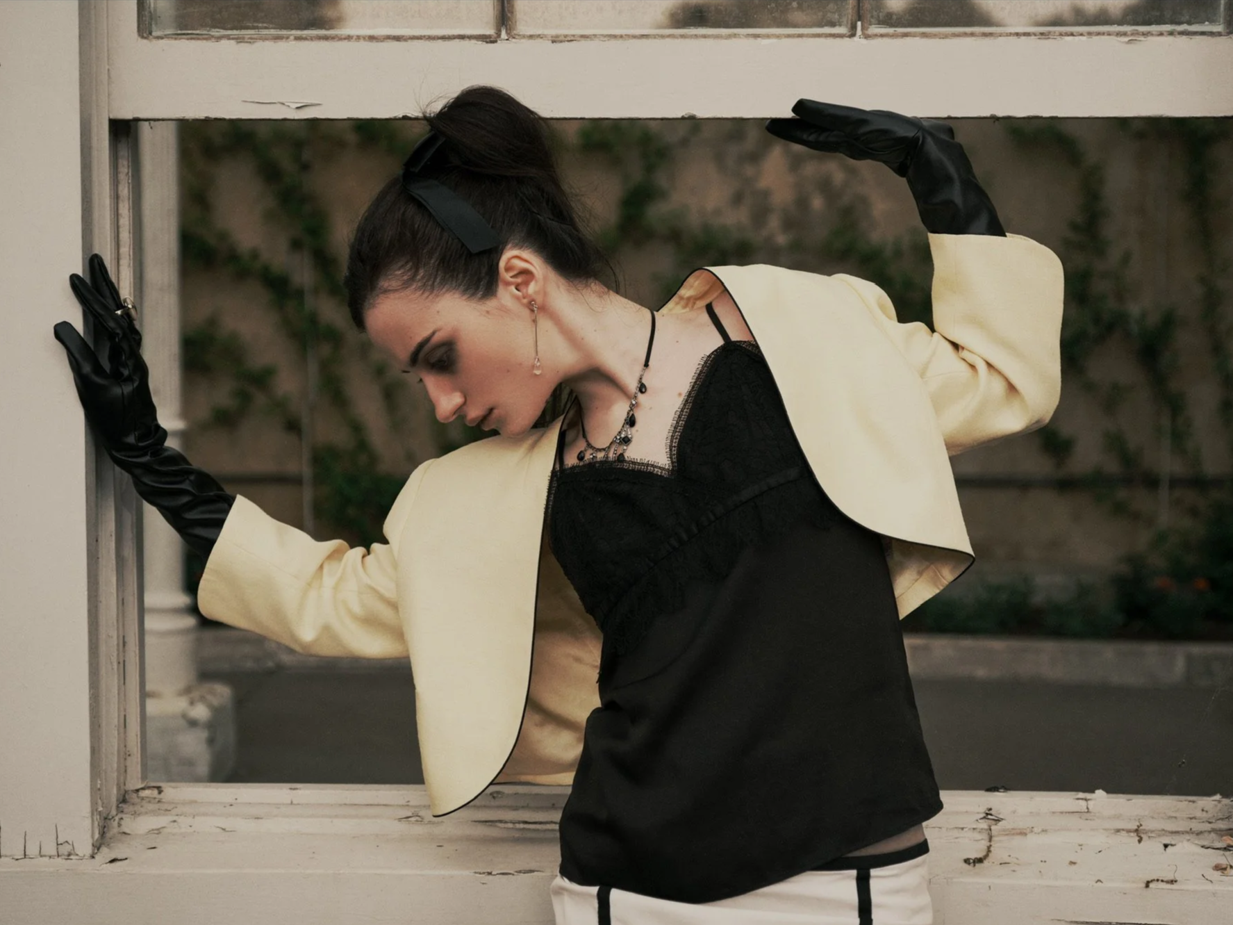 A woman wearing a cream blazer, black glove, and black dress, appears to be posing with her head tilted downward and one arm raised above a white window frame with peeling paint, standing outdoors with a blurred background.