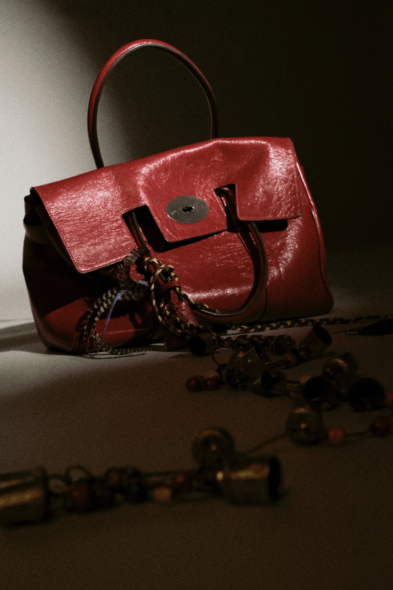 A red leather handbag with a flap closure and a circular metal detail, placed on a dark surface with a string of beads in front and a dark background.