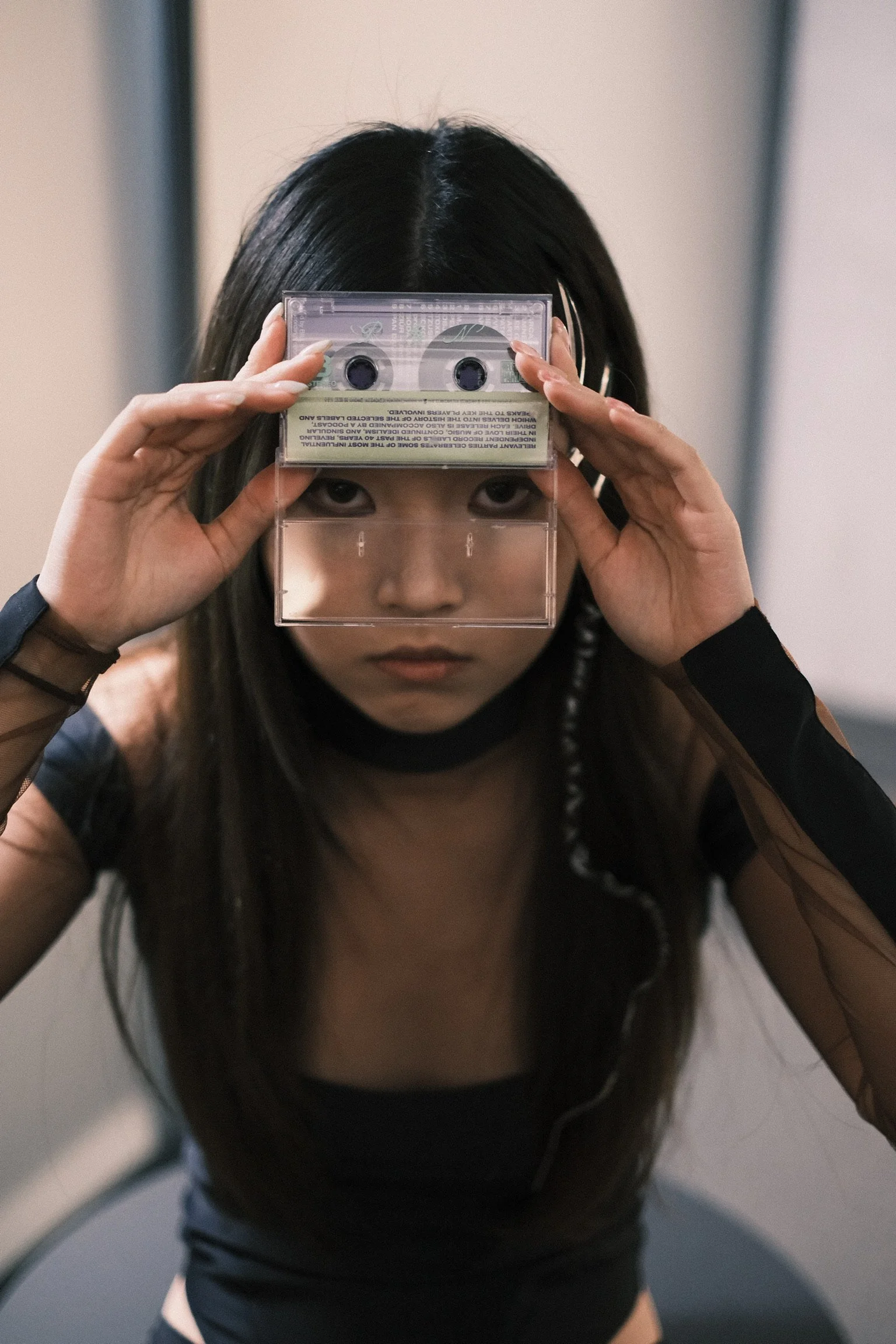 A young woman with long dark hair, wearing a black outfit and mesh sleeves, holding a cassette tape over her face, looking directly at the camera
