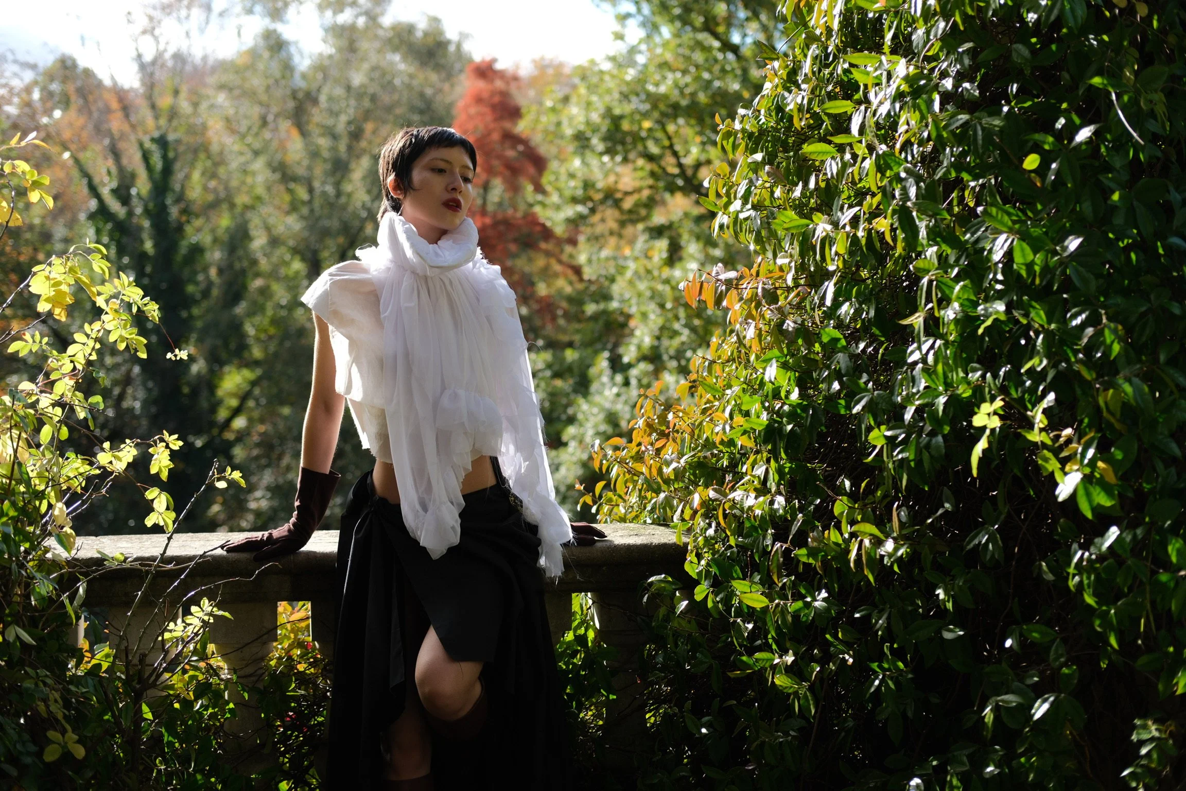 A person with short dark hair and fair skin, dressed in a white ruffled top and black skirt, sitting outdoors on a stone ledge surrounded by green bushes and trees with autumn-colored leaves.