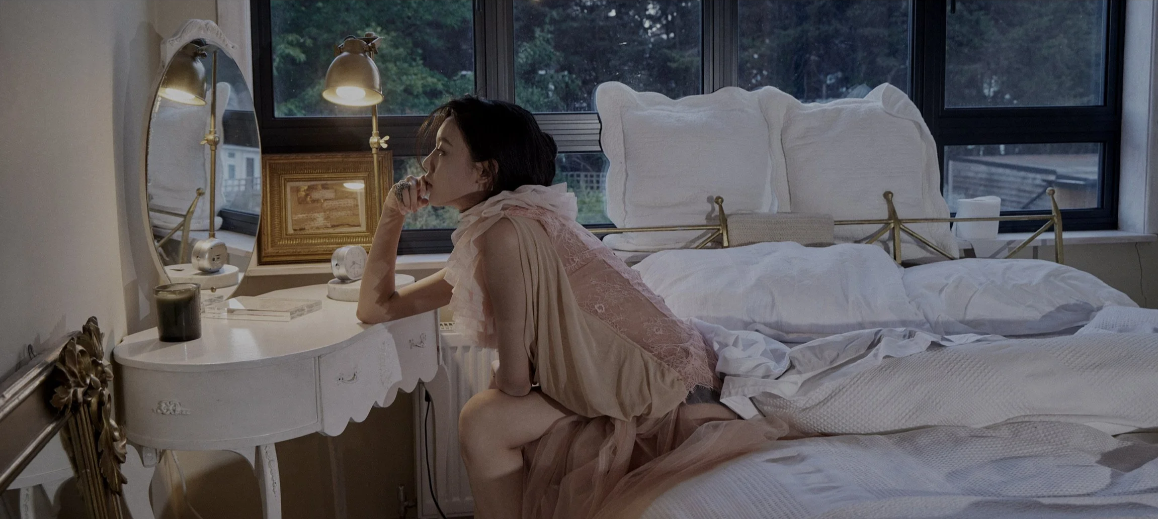 A woman with short dark hair wearing a light pink lace dress sitting on a bed, resting her chin on her hand, in a bedroom with large windows, a white bedside table, and various decorative items.