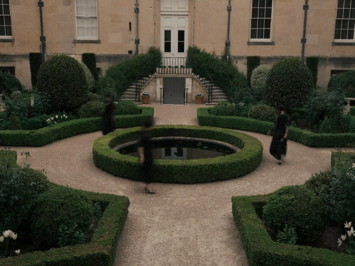 A symmetrical garden courtyard with trimmed bushes, a central circular water feature, and stairs leading to a building entrance in the background, with two blurred people walking.