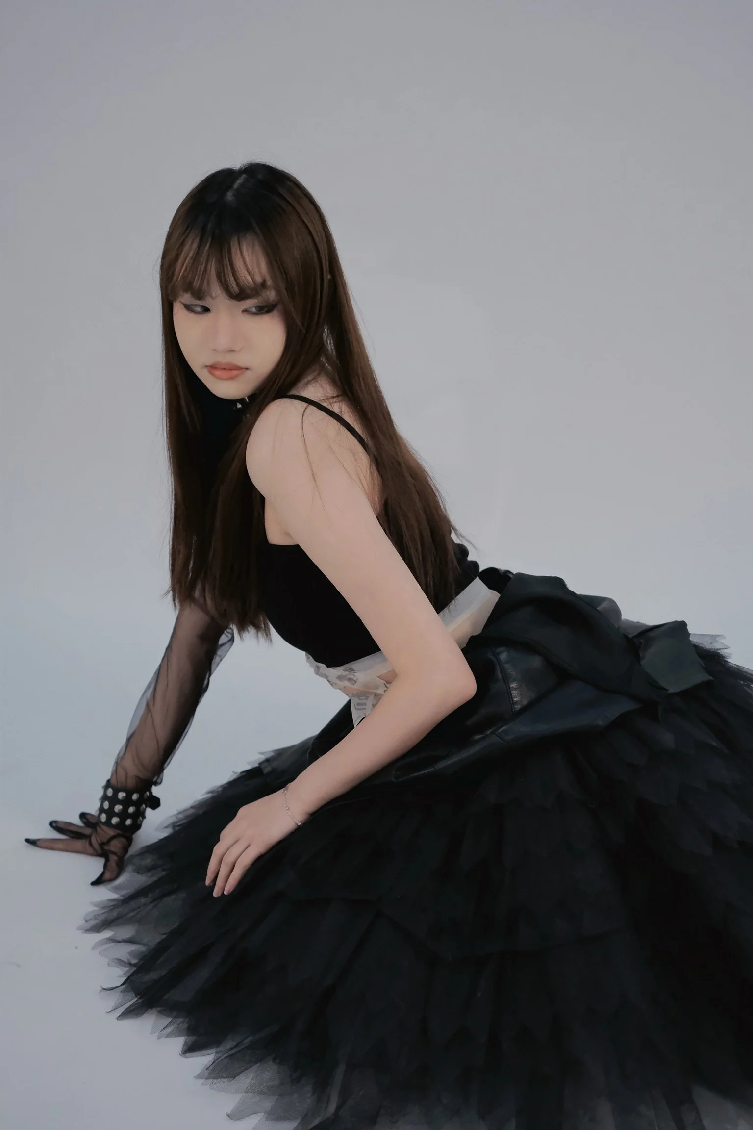 A young woman with long brown hair and bangs is wearing a black sleeveless top with sheer long sleeves, a black tulle skirt, and black accessories. She is posing on her hands and knees against a plain gray background, looking down to her left with a serious expression.