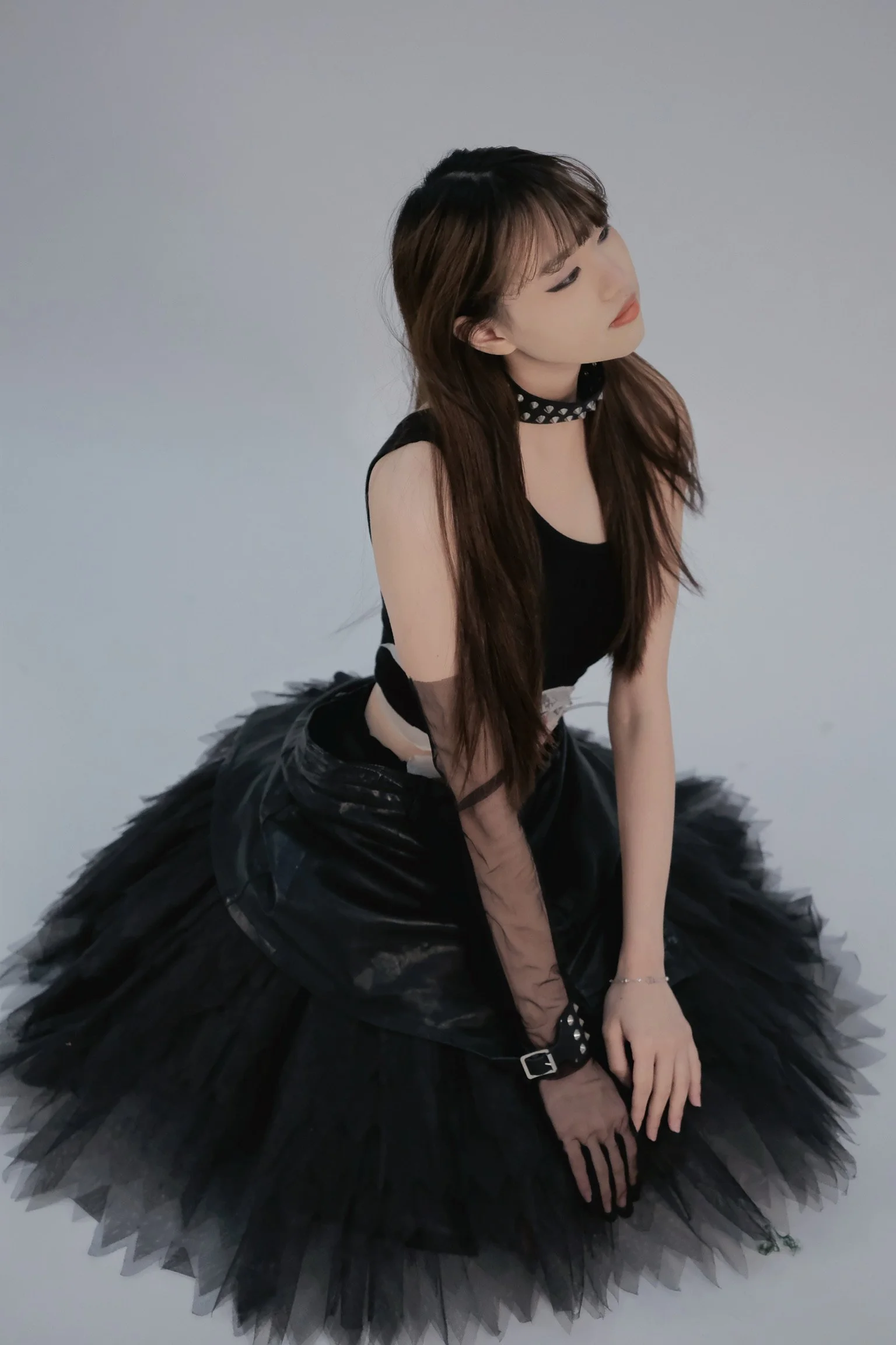 A young woman with long brown hair and bangs, wearing a black sleeveless top, a studded choker, black fingerless gloves, and a black layered tulle skirt, is sitting on the floor with her head tilted and eyes closed, against a plain light gray background.