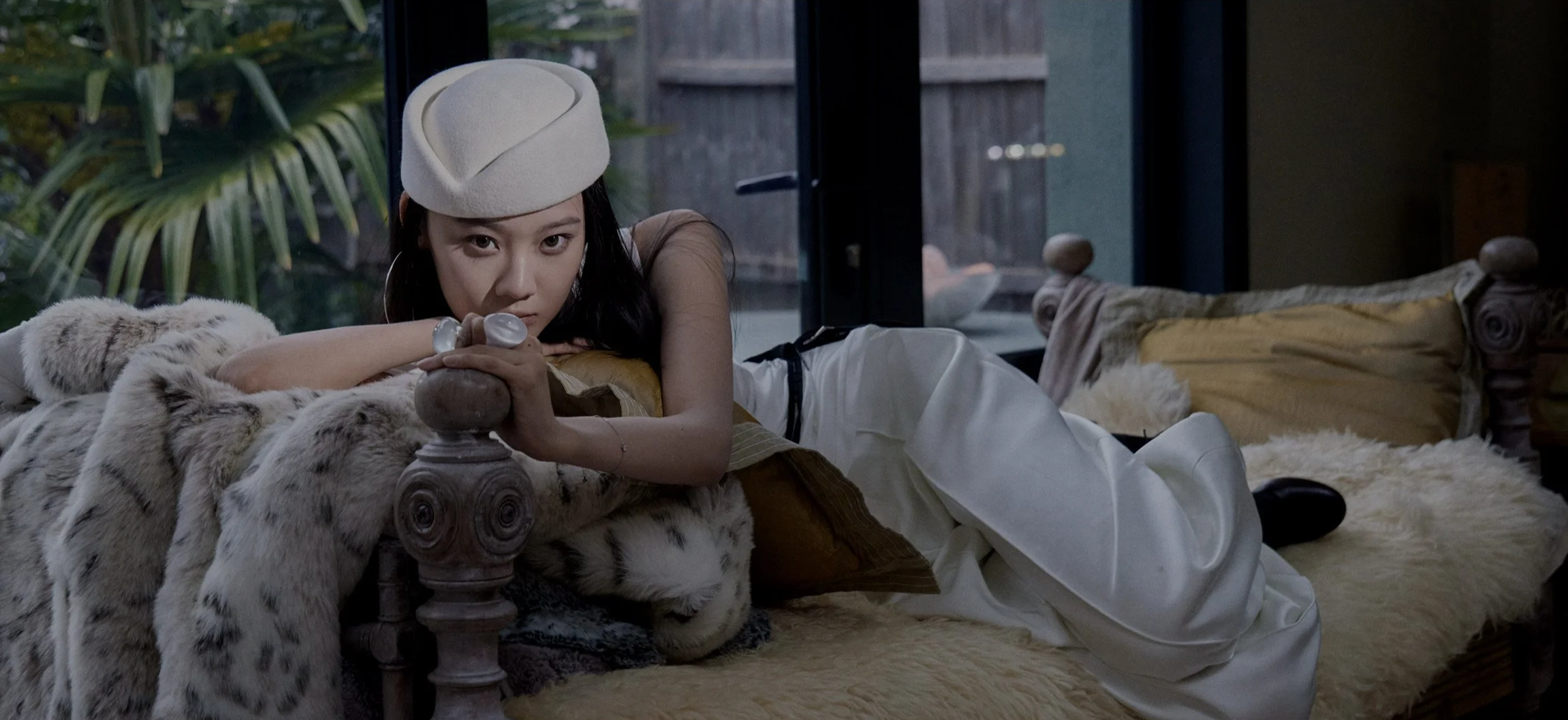A woman lying on a vintage wooden bed, resting her head on her arms with her eyes looking at the camera, wearing a white beret and white pants, in a room with a large window and greenery outside.