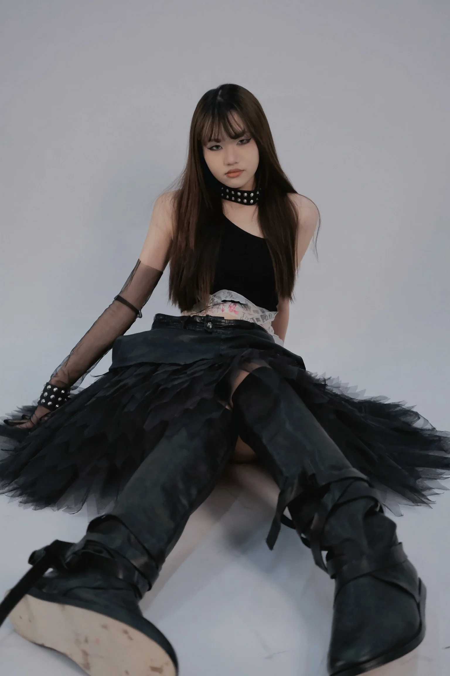 A young woman with long brown hair and makeup, wearing a black sleeveless top, black mesh sleeves, a black choker with silver studs, and black pants with shiny black boots, sitting on the ground against a plain gray background.