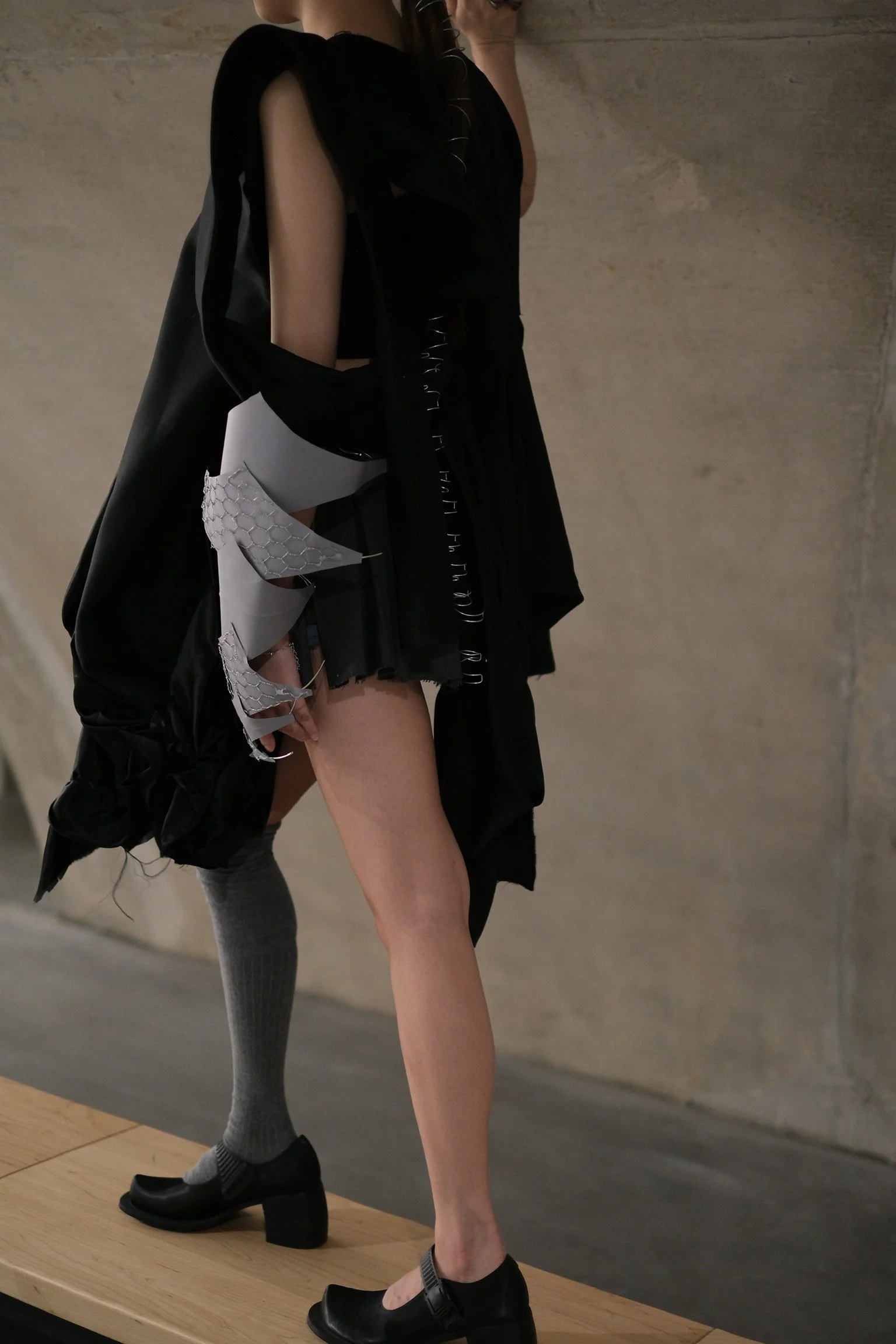 Close-up of two models wearing black footwear, featuring different clothing styles, with a focus on their legs and shoes, standing on a wooden platform against a neutral background.