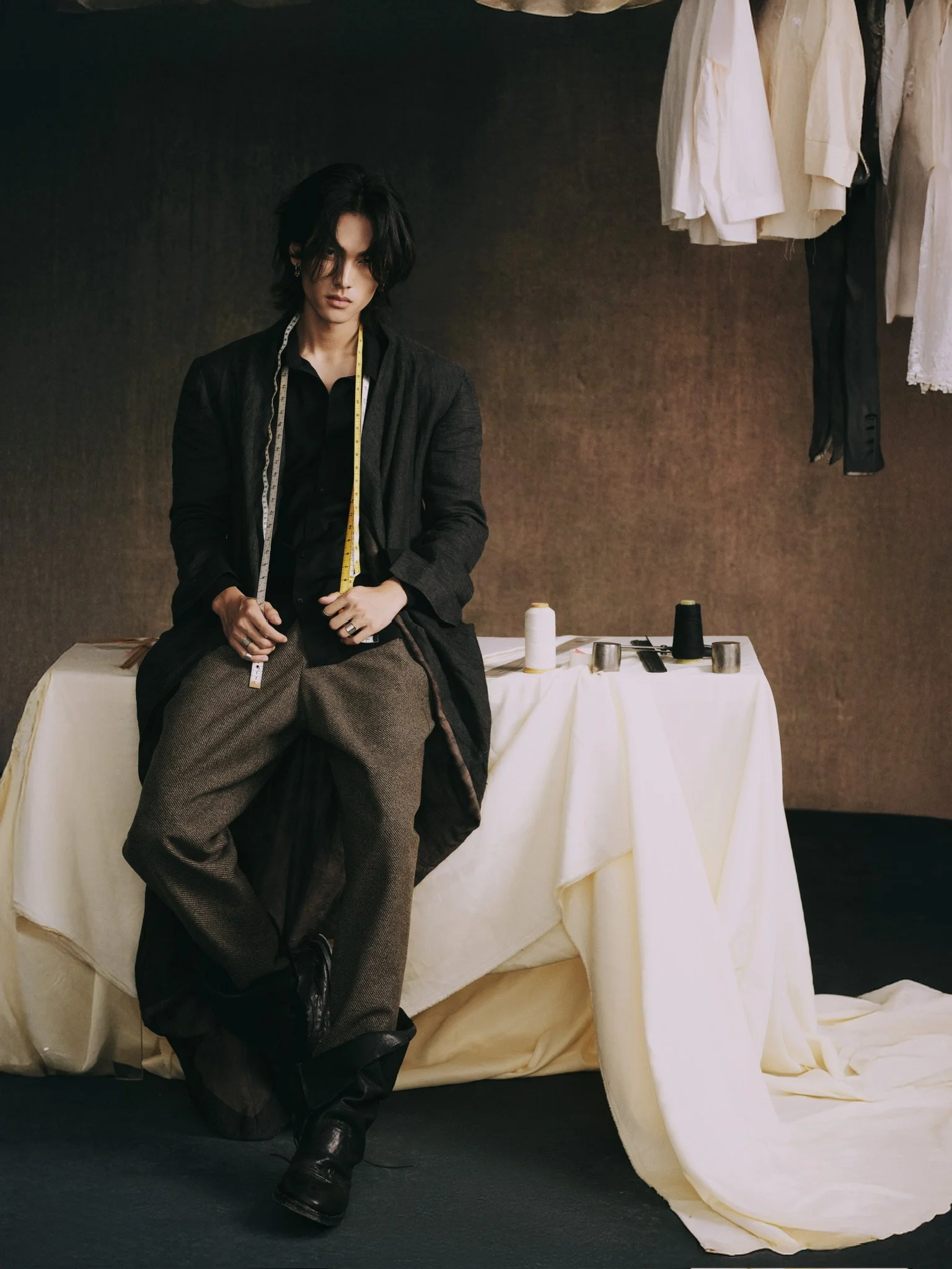 A person with dark hair sitting on a table with sewing materials, including fabric, scissors, and spools of thread. There are clothes hanging above and a cream-colored tablecloth.