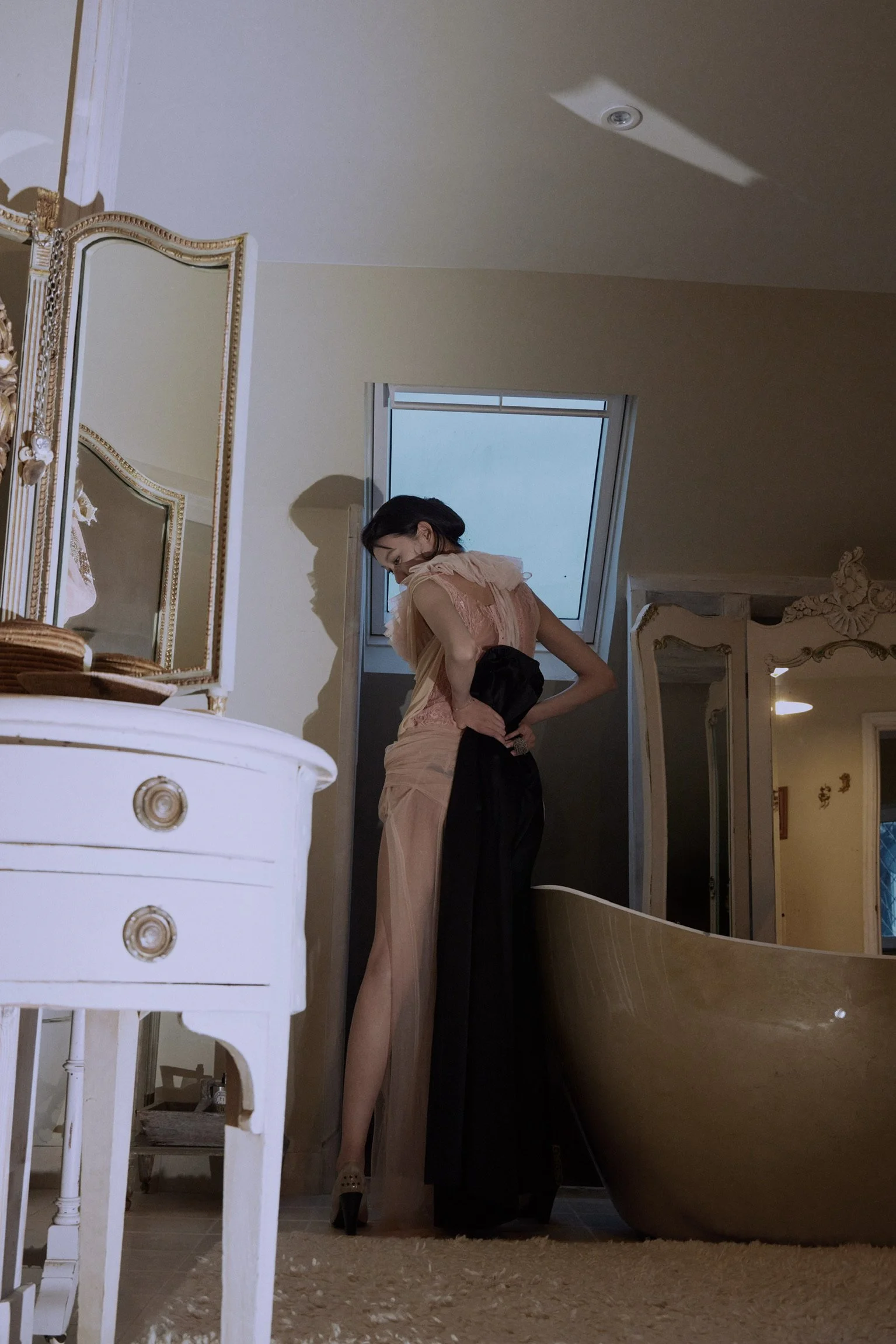A woman in a peach-colored dress and high heels is standing in a room with ornate furniture, a large bathtub, and a ceiling with a skylight. The room has a vintage aesthetic with a cream color palette.