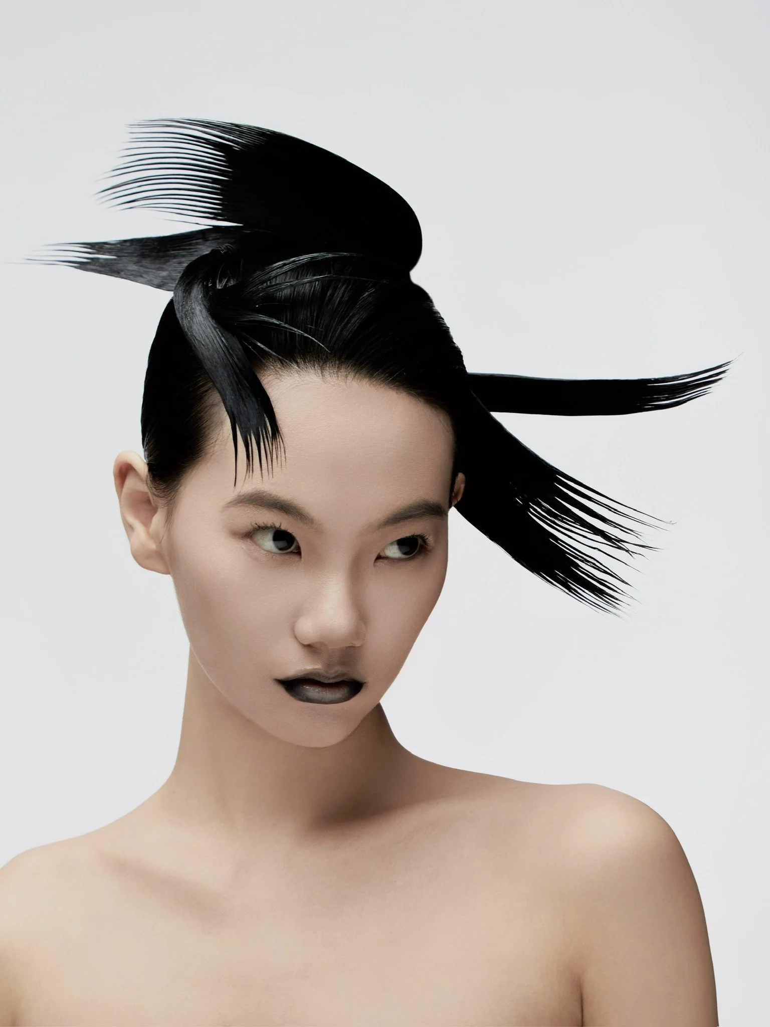 A woman with black lipstick and eye makeup, wearing an avant-garde hairstyle made of glossy black feathers or strands styled into a dramatic, sculptural shape, against a plain light gray background.
