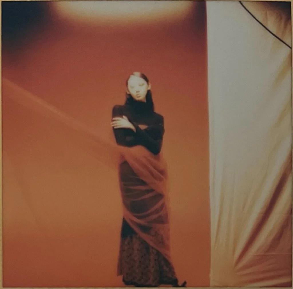 A woman in a black long-sleeve top and a long, flowing skirt standing against a warm-colored background with her arms crossed.