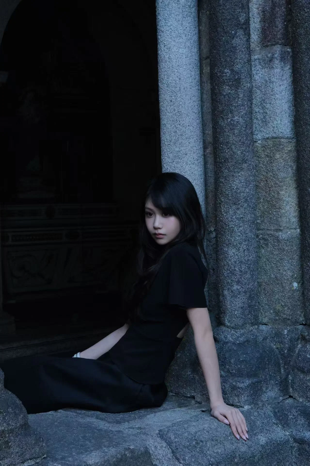 A young woman with black hair and pale skin leaning against a stone wall outdoors, wearing a black dress, with her right arm resting on the ground and her left arm bent, looking at the camera with a neutral expression.
