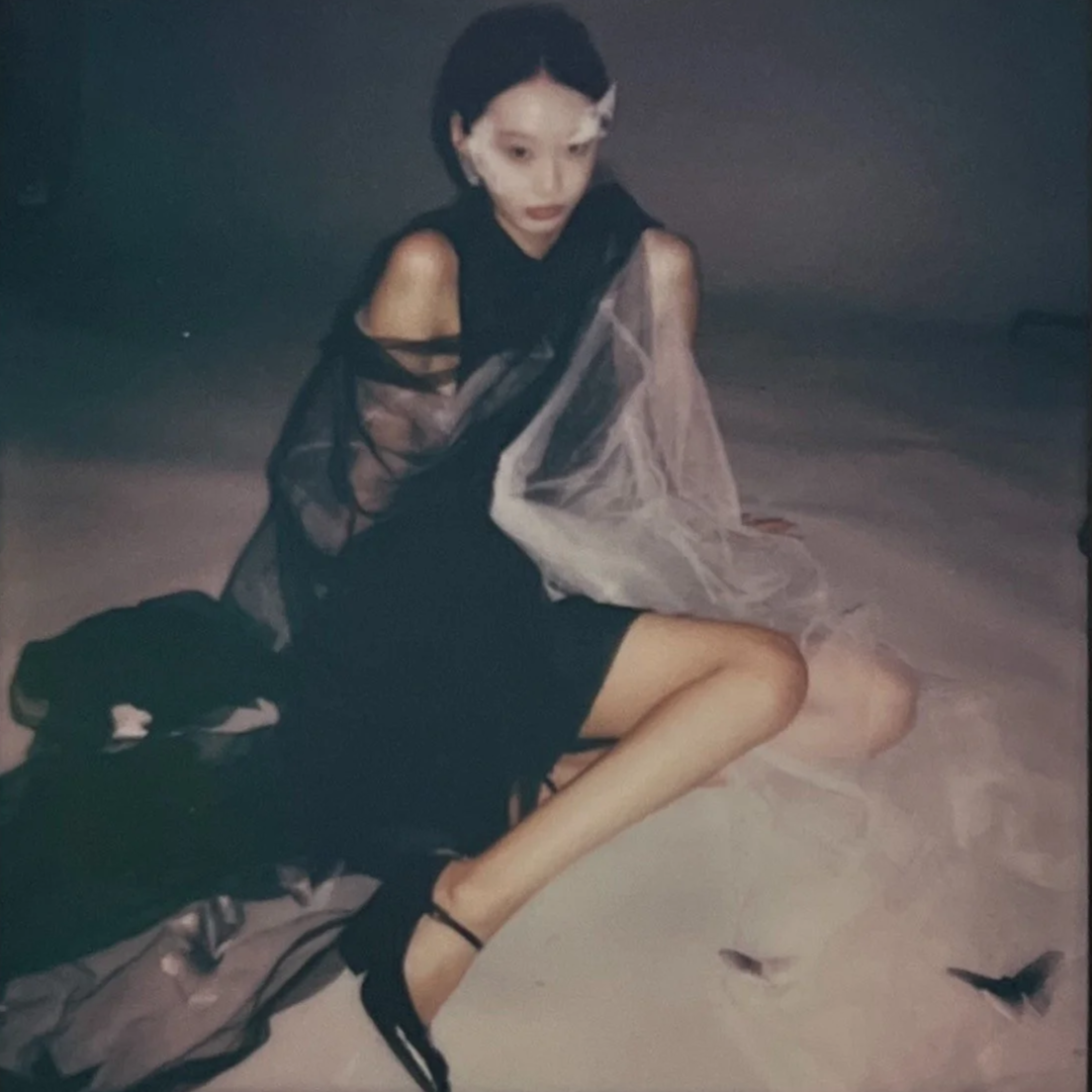 A young woman sitting on the floor in a black dress with sheer fabric sleeves, wearing black heels, with a sheer cape or shawl draped over her shoulders, in a dimly lit environment.
